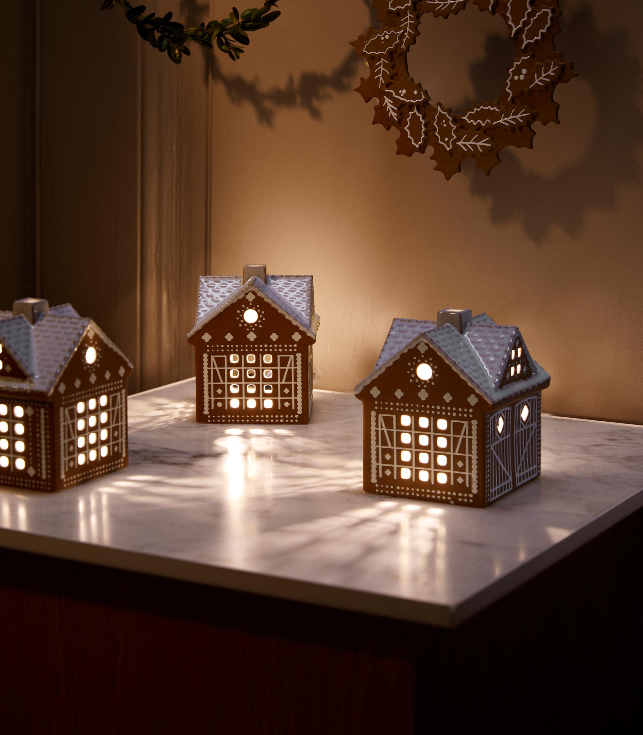 3 ljuslyktor i form av pepparkakshus som står på ett bord och lyser upp ett mörkt rum