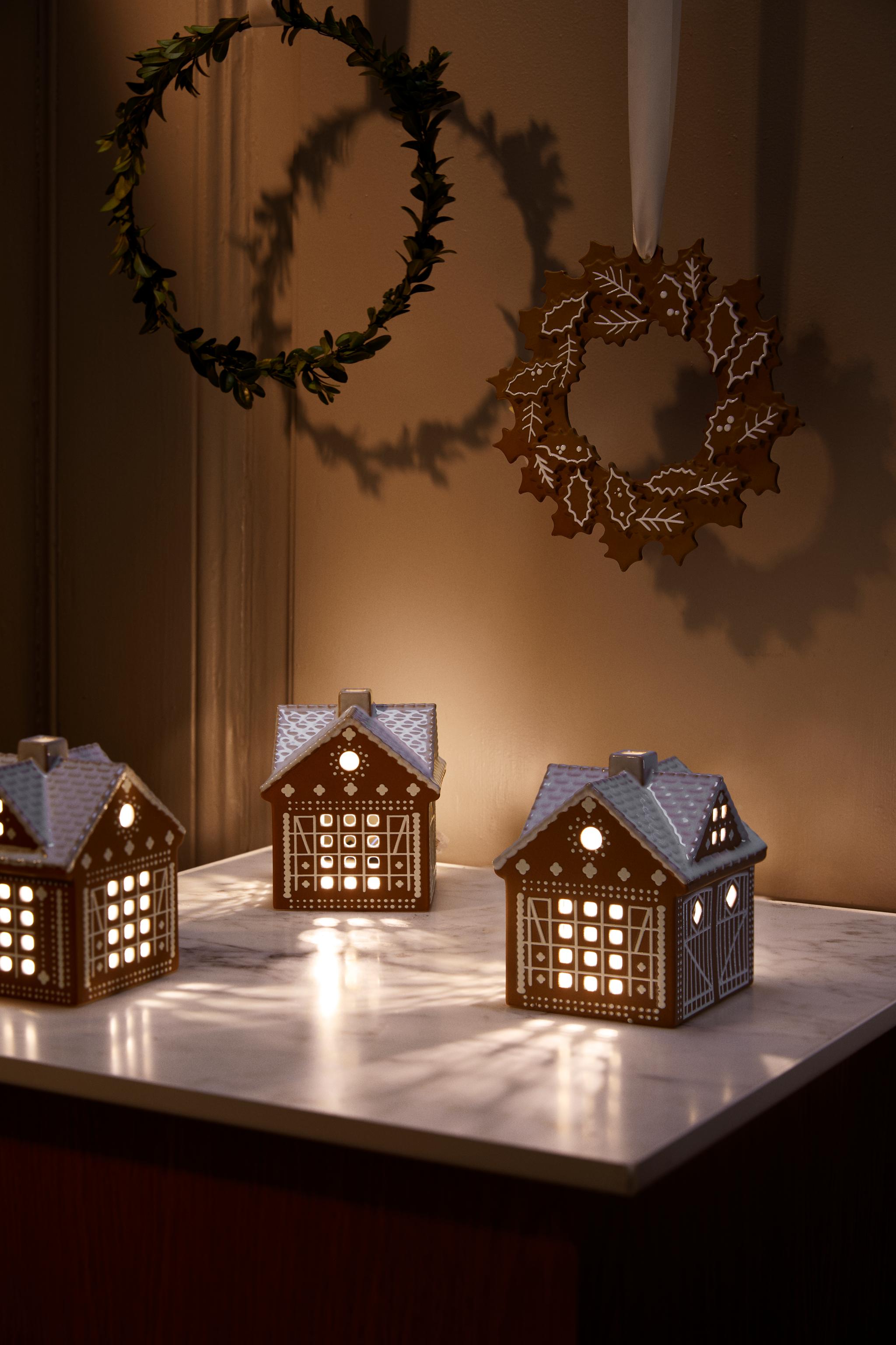 Kolme hehkuvaa piparkakkutalolyhtyä marmoritason päällä, ja kaksi jouluista kranssia roikkuu seinällä.