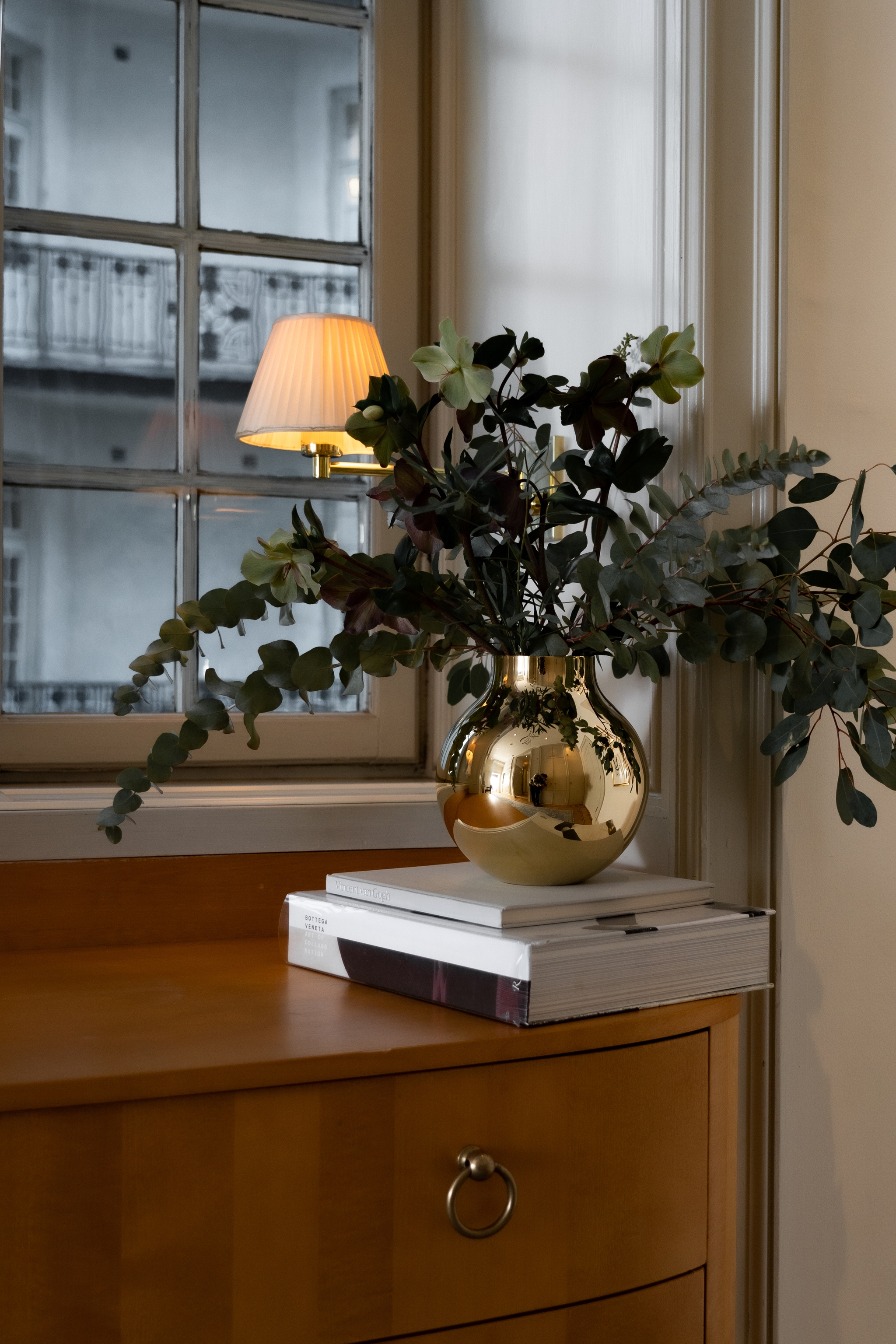 Blomsterarrangemang i en blank guldfärgad vas på böcker framför ett fönster och en tänd vägglampa.