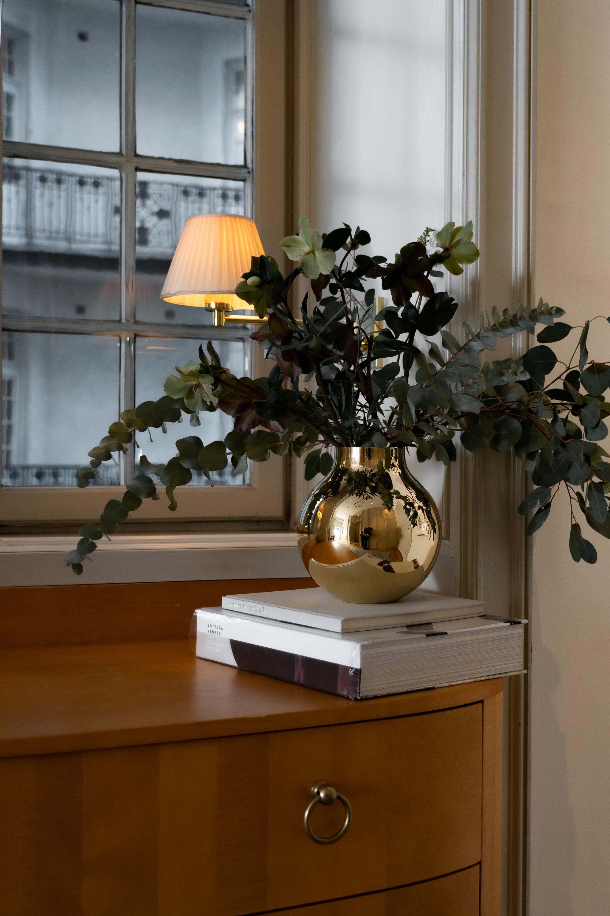 Blomsterarrangemang i en blank guldfärgad vas på böcker framför ett fönster och en tänd vägglampa.
