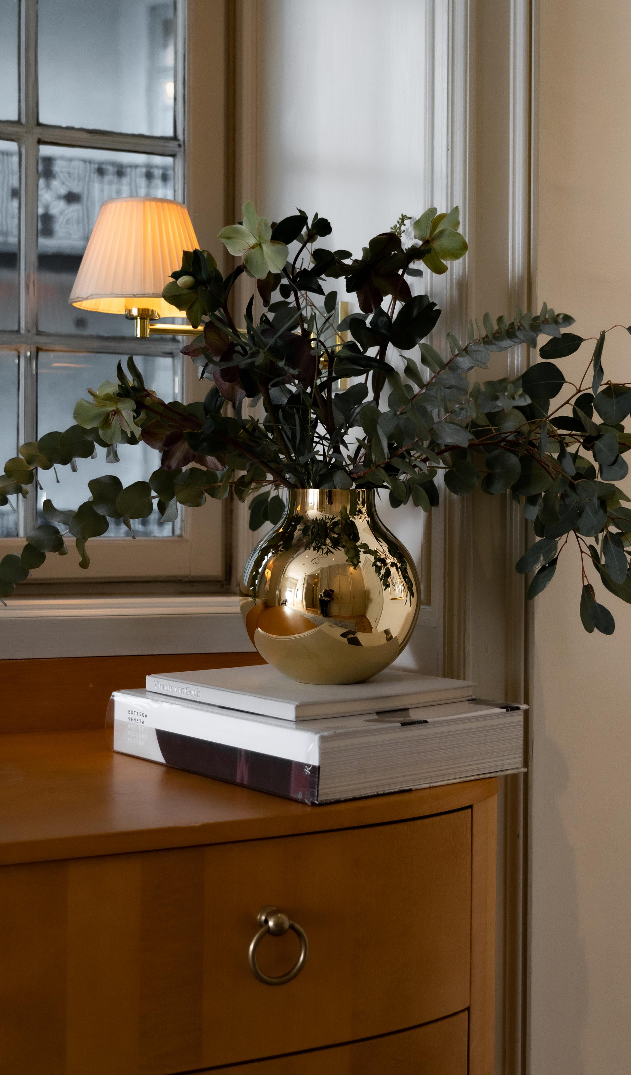 Blomsterarrangemang i en blank guldfärgad vas på böcker framför ett fönster och en tänd vägglampa.