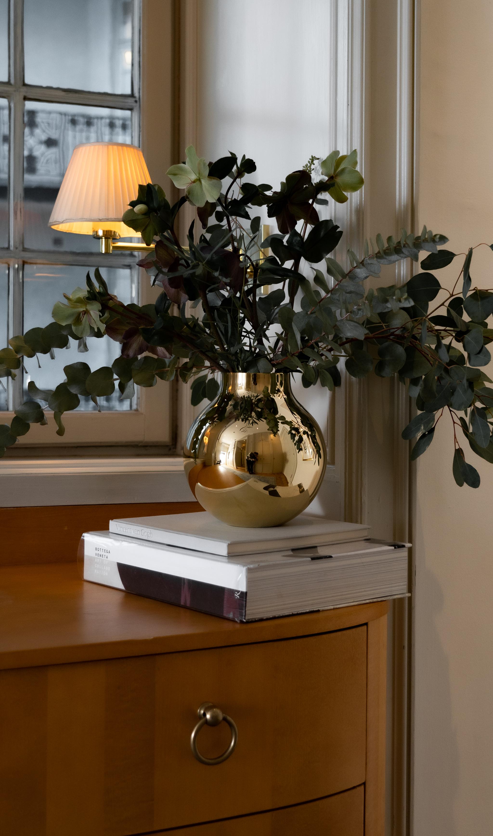 Blomster og eukalyptus i en blank gullvase på bøker ved et vindu og en tent lampe.