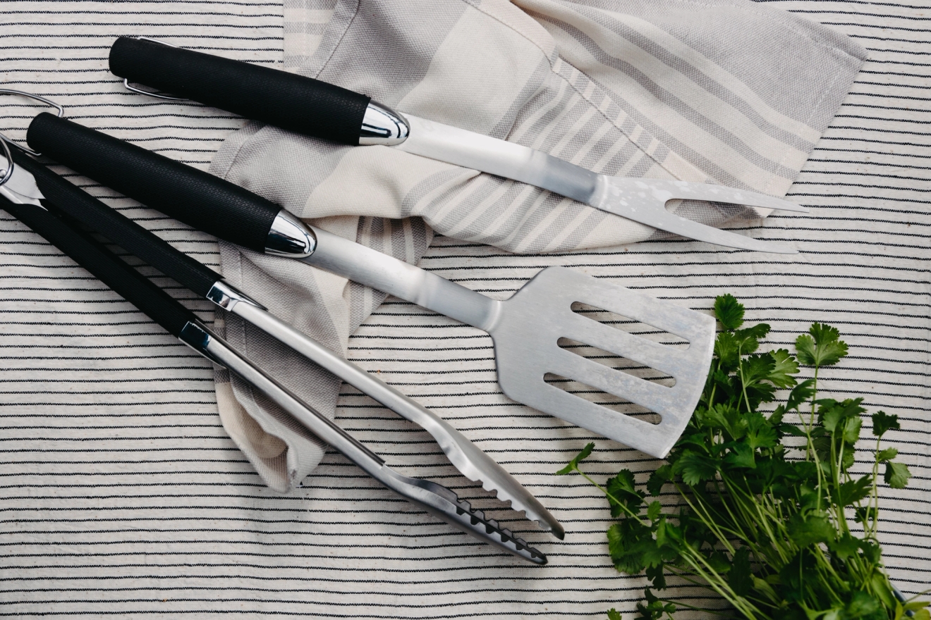 En uppsättning grillredskap med svarta skaft (tång, stekspade och gaffel) och en knippa persilja på en randig duk.