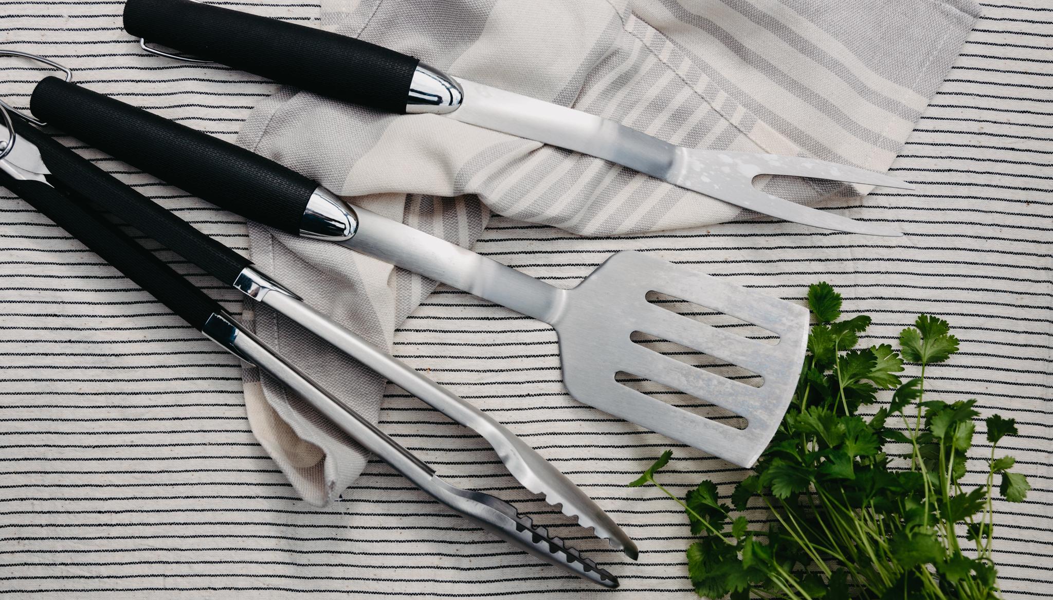 Kolme grillausvälinettä – pihdit, lasta ja paistohaarukka – raidallisen pyyhkeen päällä tuoretta persiljaa.