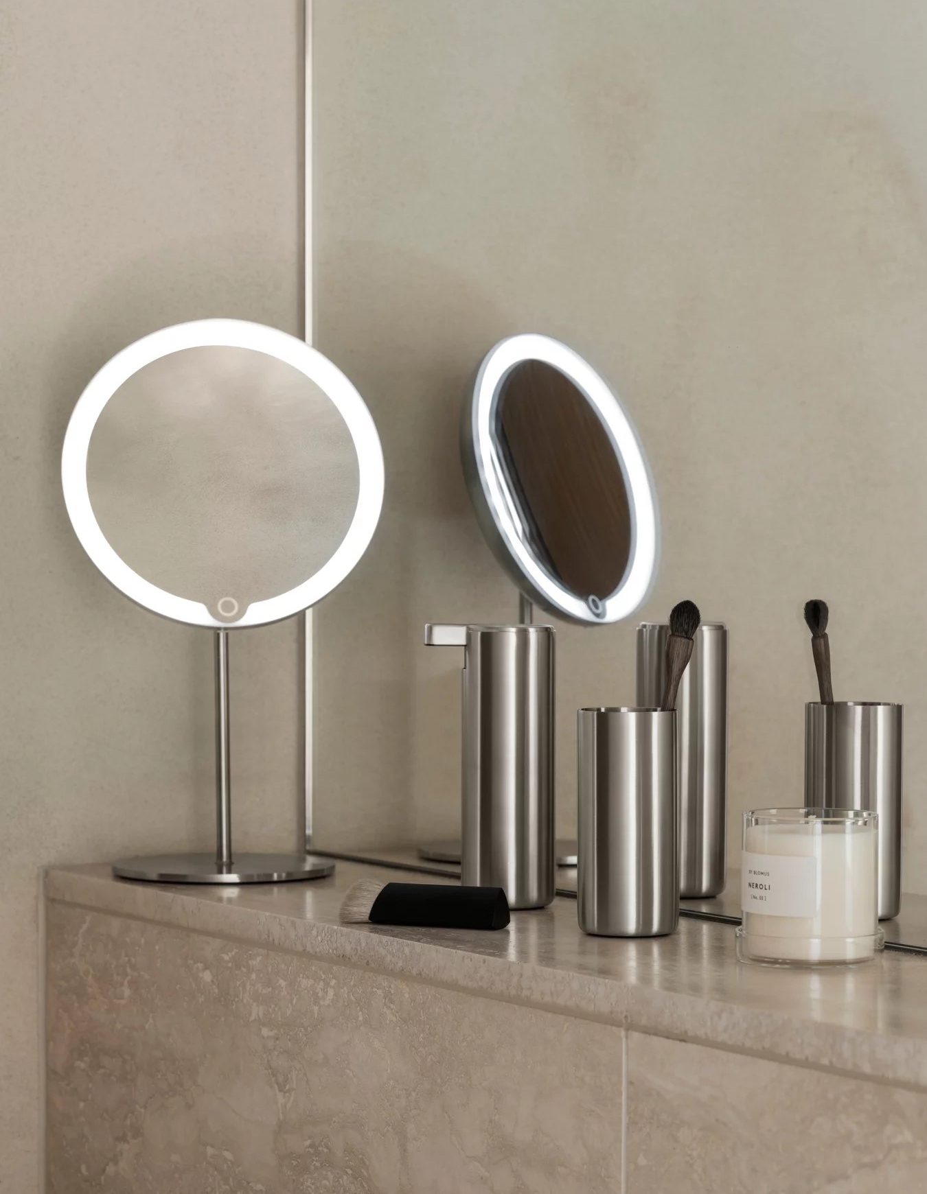 Rund sminkspegel med LED-belysning, sminkborstar och doftljus på en elegant badrumsbänk.