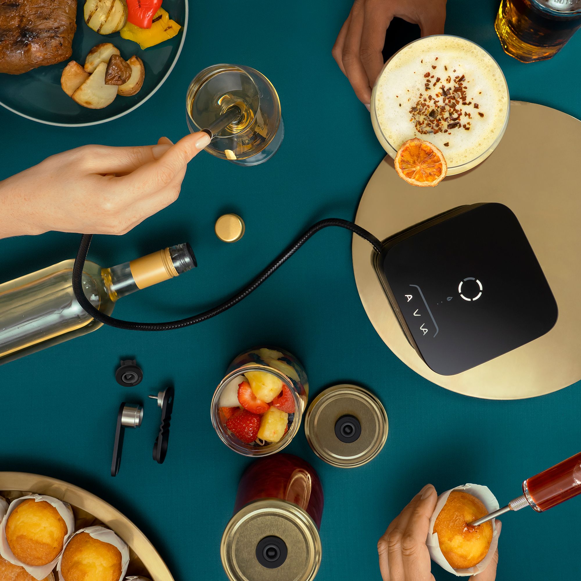 Overhead view of an AVA infuser device connected to a wine bottle and a wine glass, surrounded by hands, grilled food, cocktails, and muffins being injected with flavor on a dark teal table.