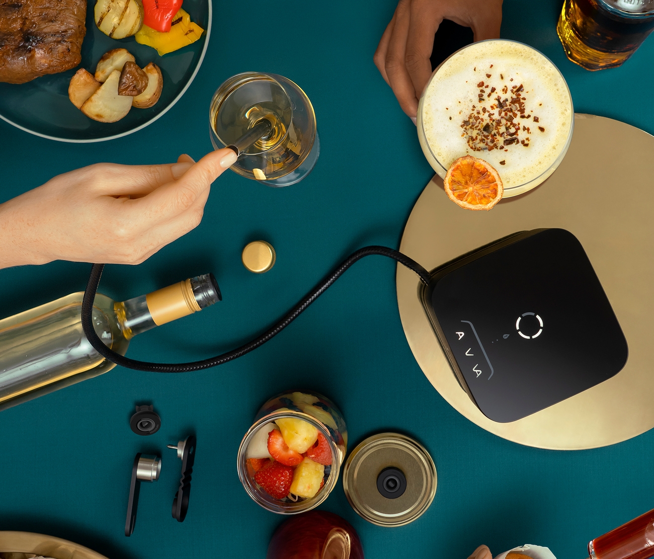 Ylhäältä kuvattu näkymä AVA-infusointilaitteesta, joka on liitetty viinipulloon ja viinilasiin. Laitteen ympärillä näkyy käsiä, grillattua ruokaa, cocktaileja sekä muffineja, joihin lisätään makua, tumman petroolinsävyisellä pöydällä.