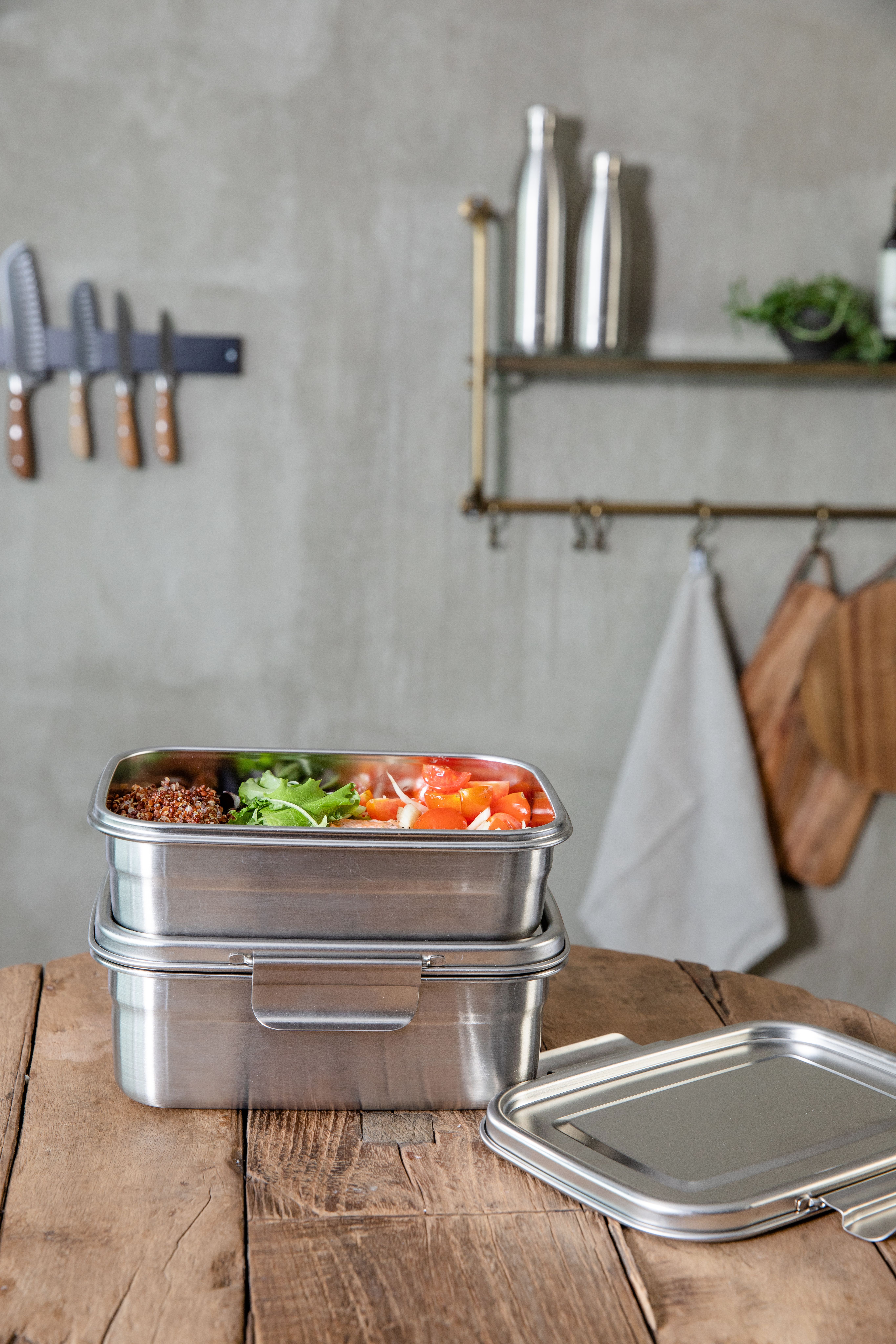 Pinottuja ruostumattomasta teräksestä valmistettuja lounasrasioita puupöydällä, yksi täynnä kvinoaa, salaattia ja tomaatteja.