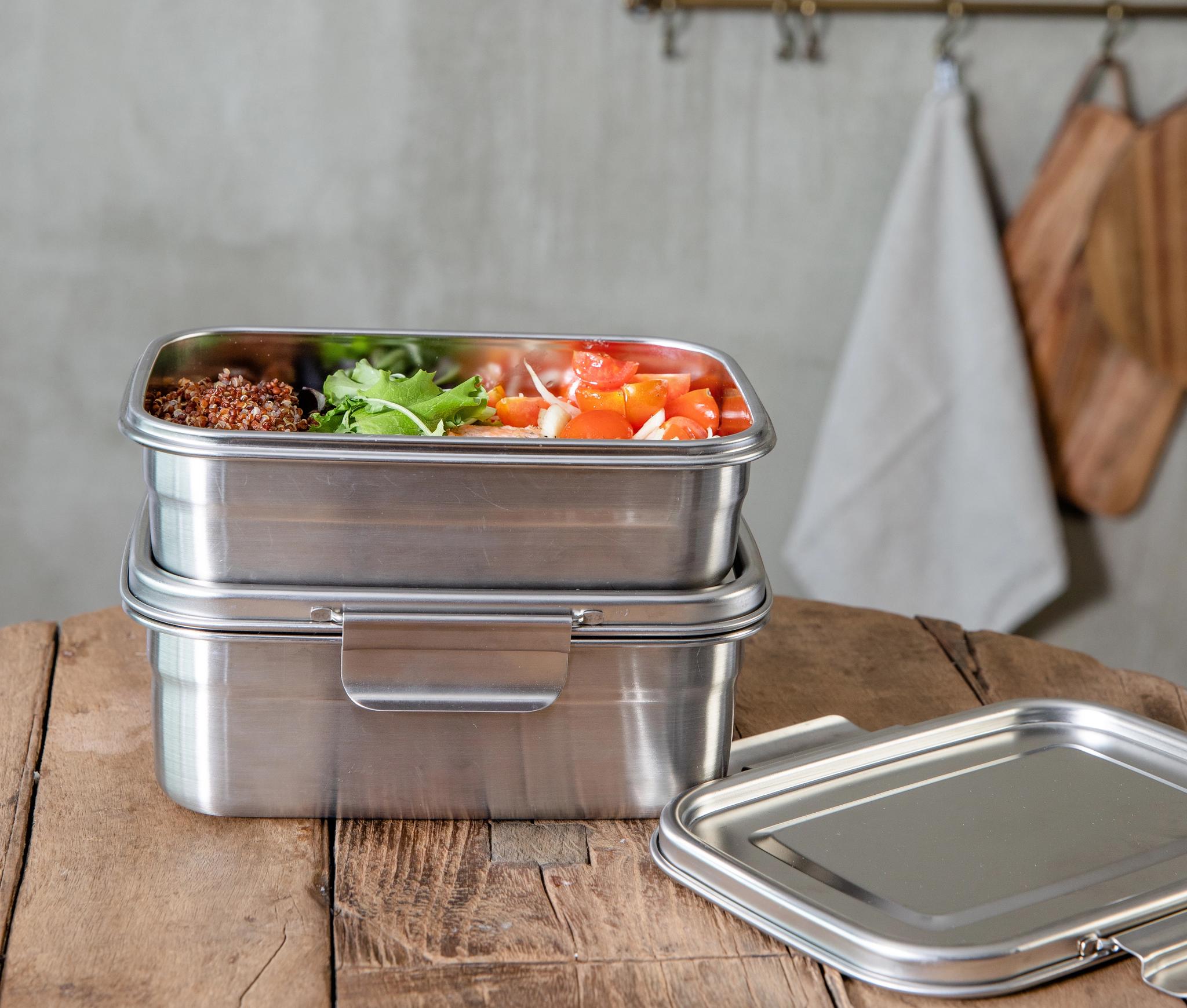 Pinottuja ruostumattomasta teräksestä valmistettuja lounasrasioita puupöydällä, yksi täynnä kvinoaa, salaattia ja tomaatteja.