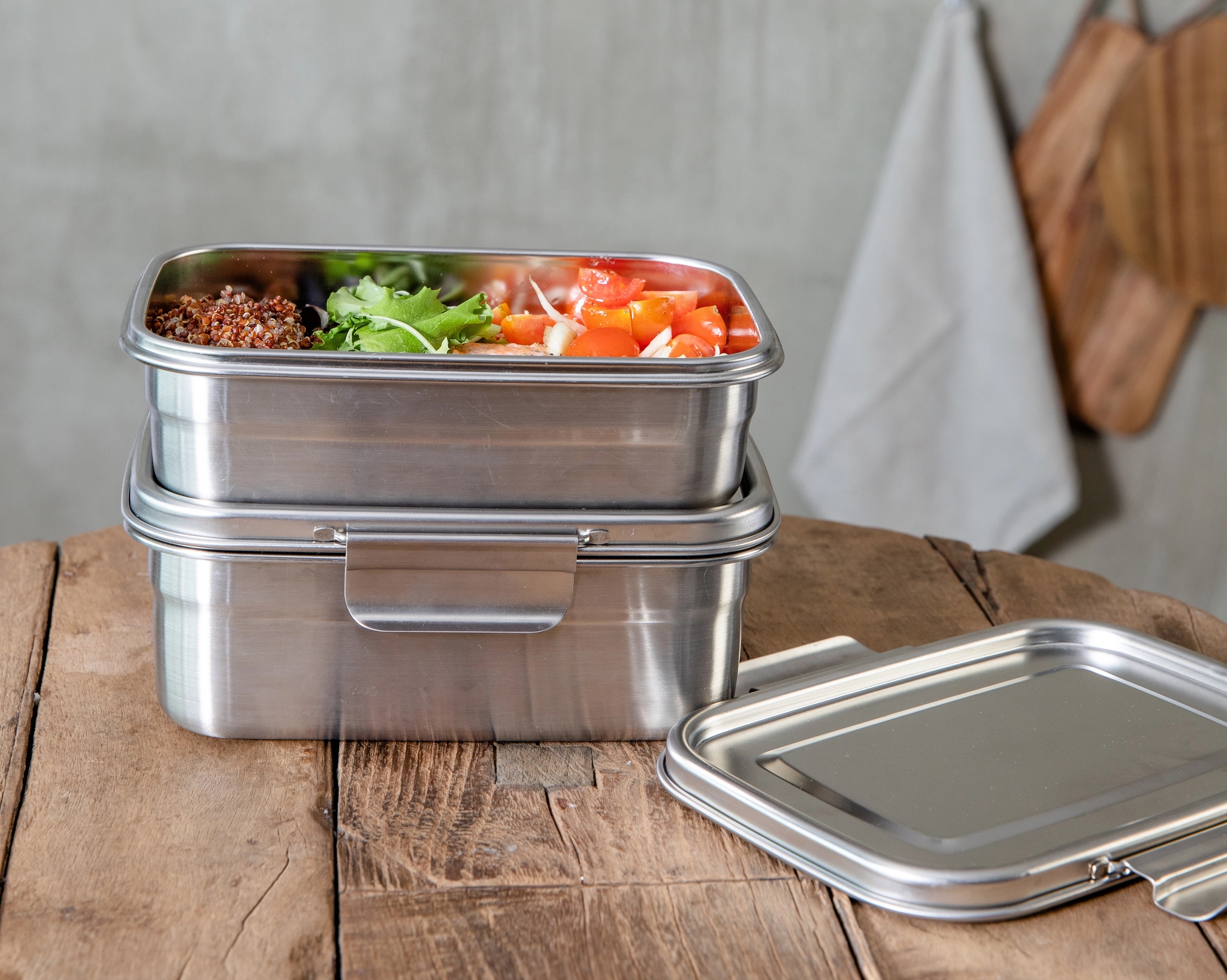 Pinottuja ruostumattomasta teräksestä valmistettuja lounasrasioita puupöydällä, yksi täynnä kvinoaa, salaattia ja tomaatteja.