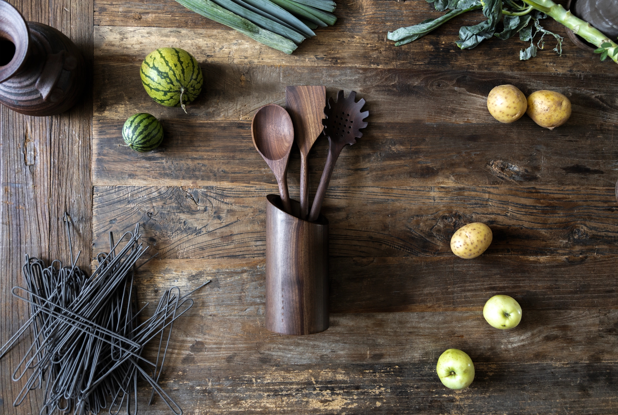 Köksredskap i trä, minivattenmeloner, potatis, gröna äpplen, gröna blad och svart pasta arrangerade på ett rustikt träbord.