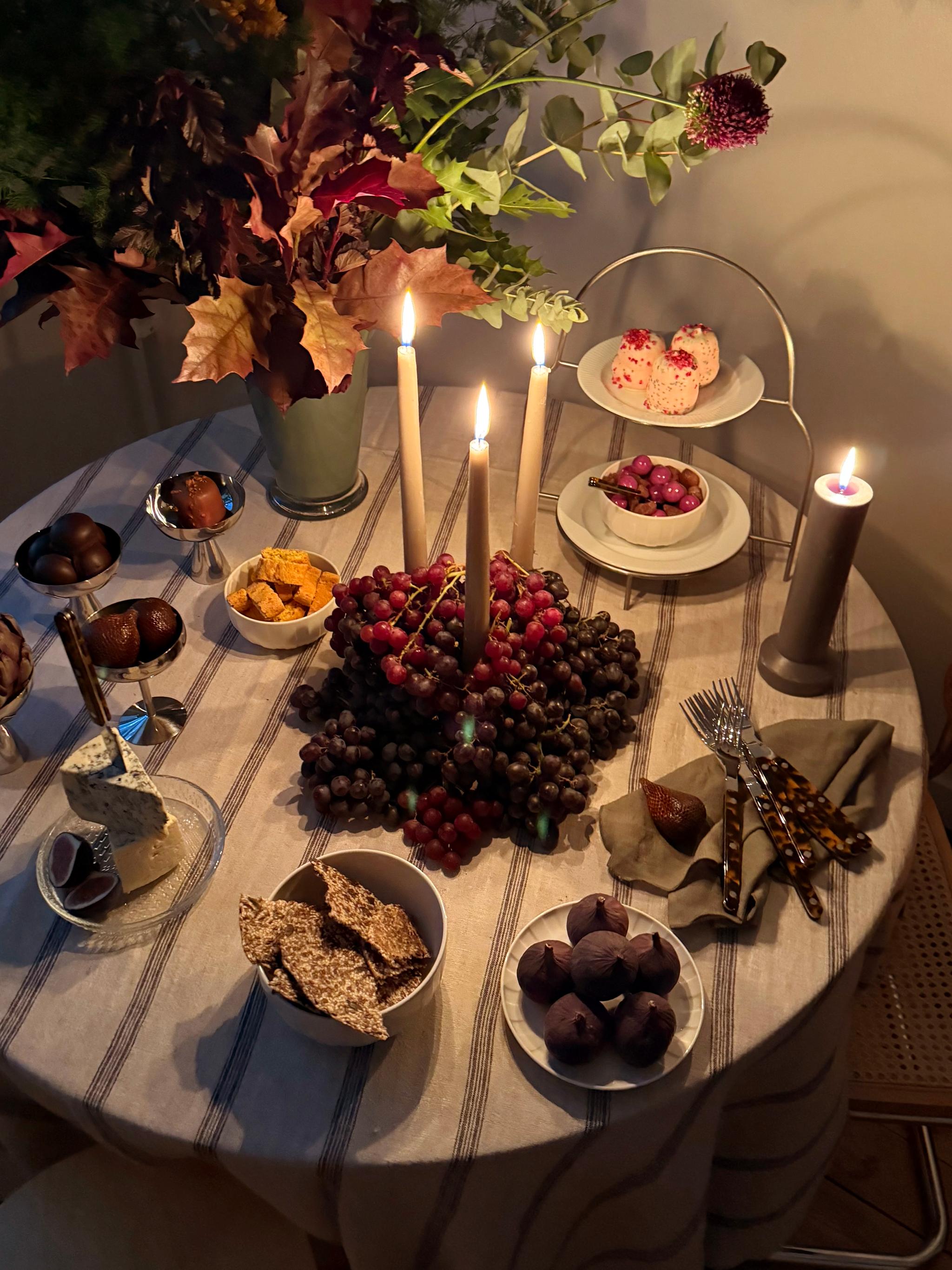 A round table set with lit candles, a large centerpiece of red and dark grapes, cheeses, figs, crackers, desserts, and an autumn floral arrangement, creating a warm, inviting spread.