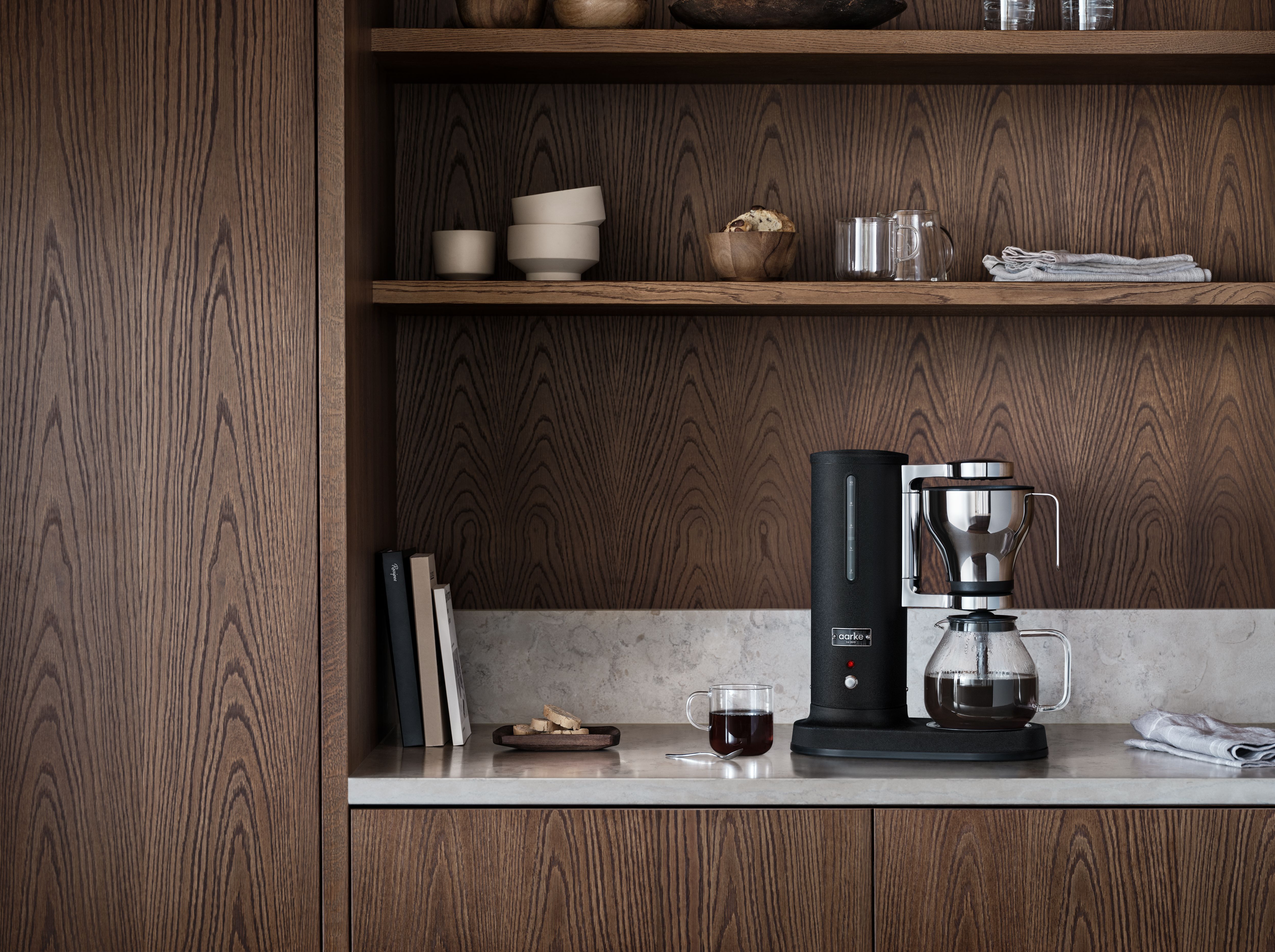 Musta kahvinkeitin ja kuppi kahvia vaalealla työtasolla keittiössä, jossa on tummat puukaapit.