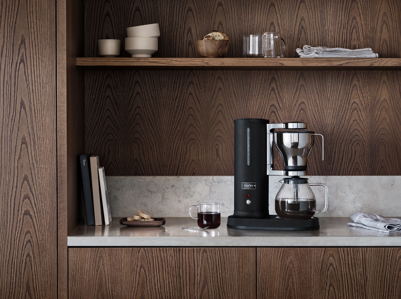 A black coffee maker and a cup of coffee on a light gray kitchen counter, surrounded by dark wood cabinets and shelves.