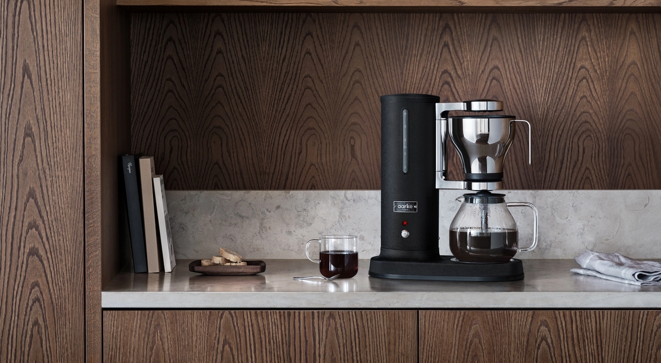 En svart kaffetrakter og en kopp kaffe på en lys benk i et kjøkken med mørke treskap.