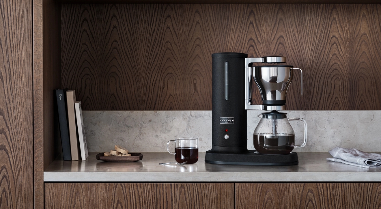 Musta kahvinkeitin ja kuppi kahvia vaalealla työtasolla keittiössä, jossa on tummat puukaapit.