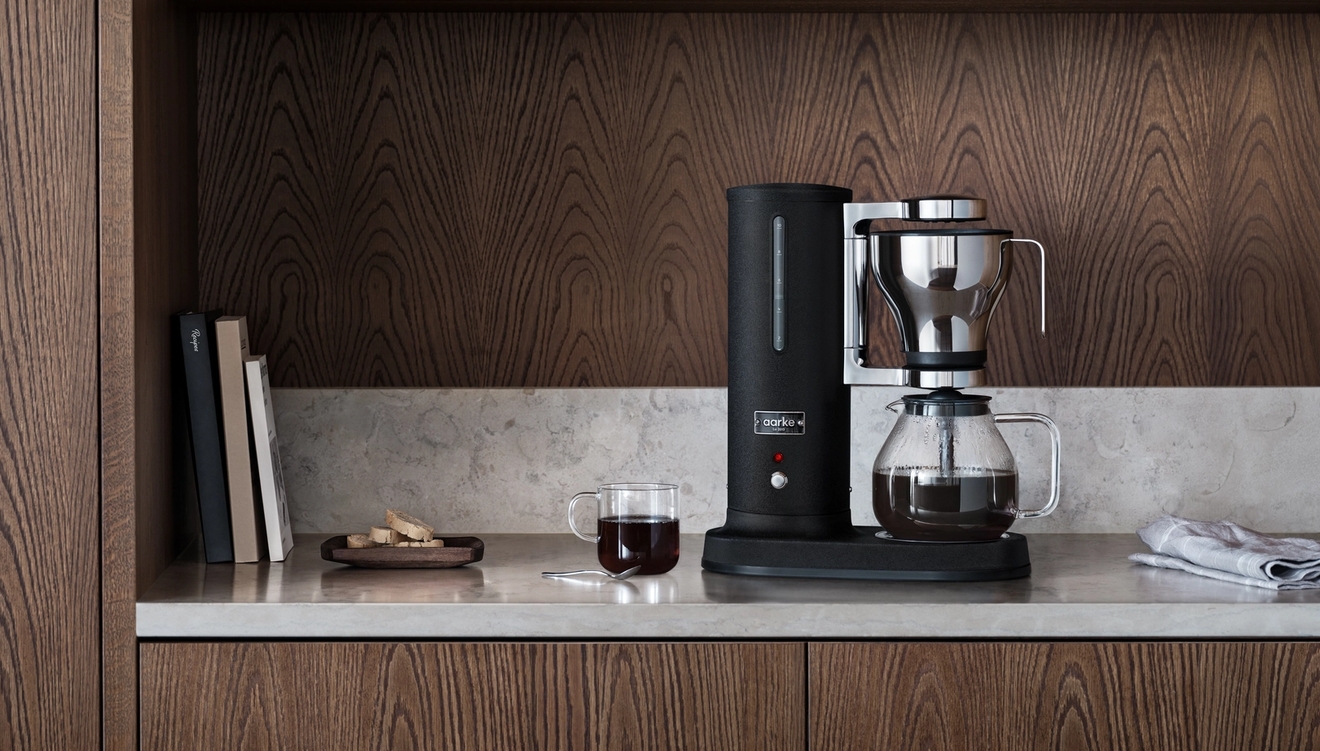 En svart kaffebryggare och en kopp kaffe på en ljus bänkskiva i ett kök med mörka träskåp.