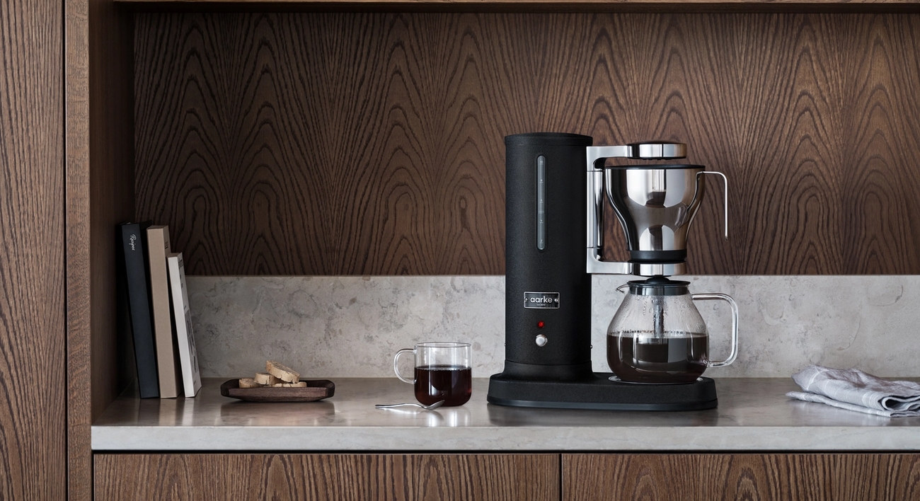 En svart kaffebryggare och en kopp kaffe på en ljus bänkskiva i ett kök med mörka träskåp.