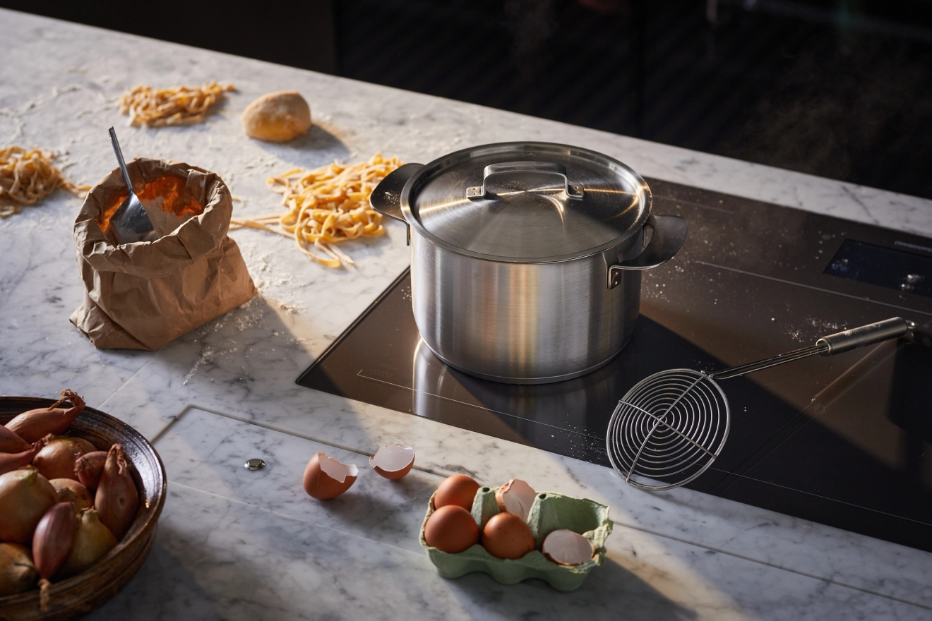 Tuorepastaa, jauhoja, kananmunia ja salottisipuleita on levitetty marmorisen keittiötason päälle kattilan viereen induktioliedellä.