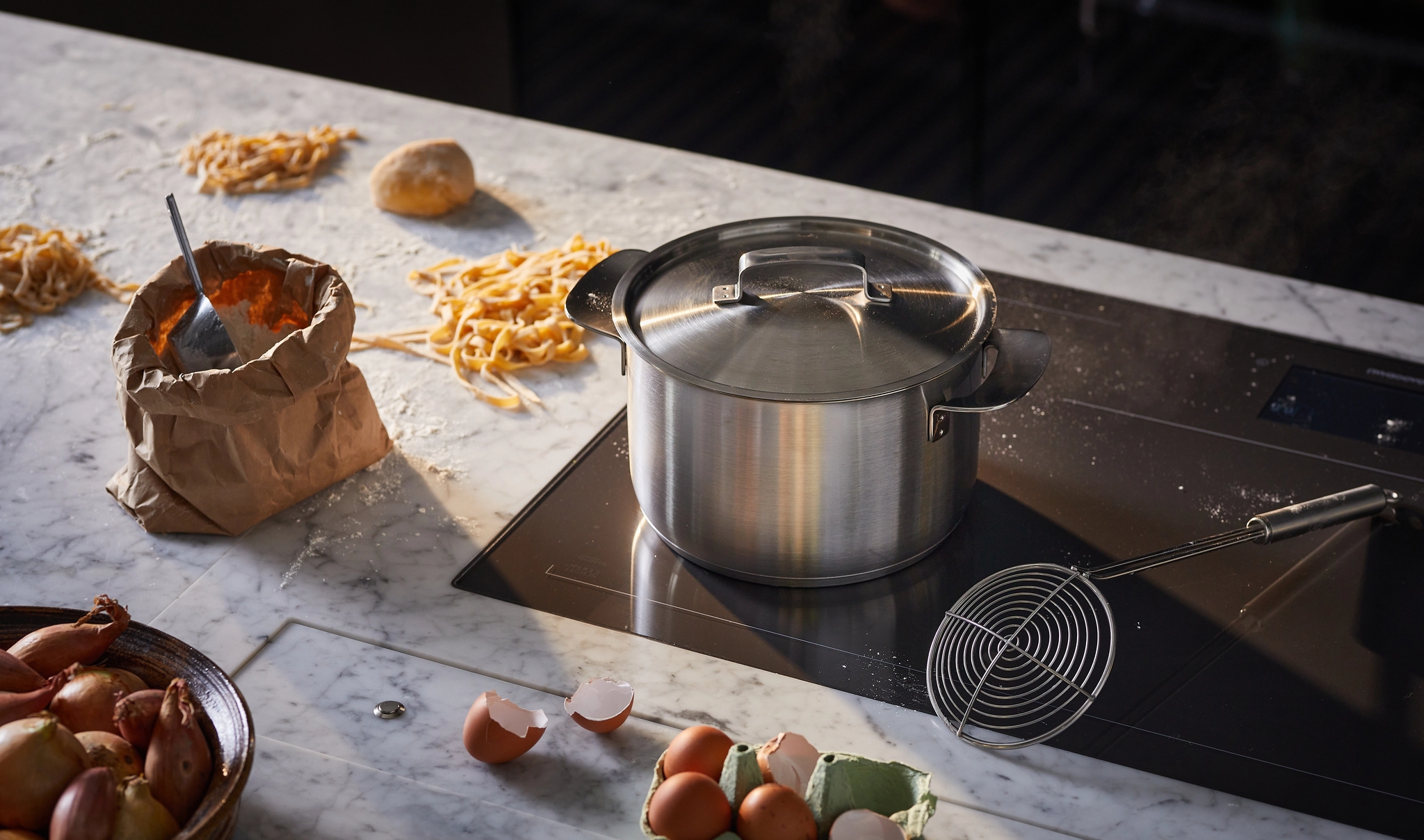 A kitchen counter covered in flour and fresh pasta strands, with a paper bag of ingredients, a bowl of shallots, and eggs next to a pot on an induction cooktop.