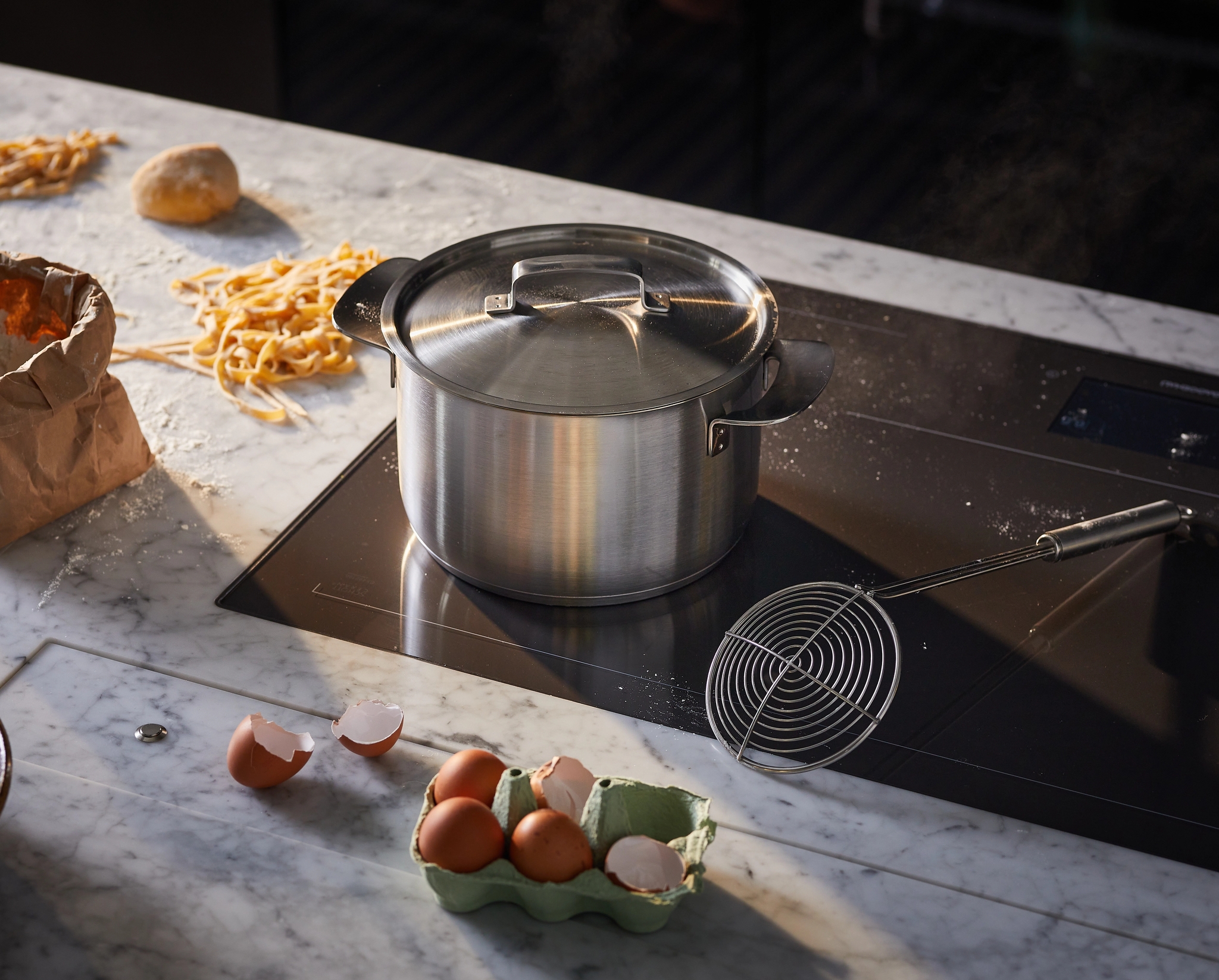 Kotitekoisen pastan ainekset hajallaan marmorisella keittiötasolla, kattila induktioliesitasolla.