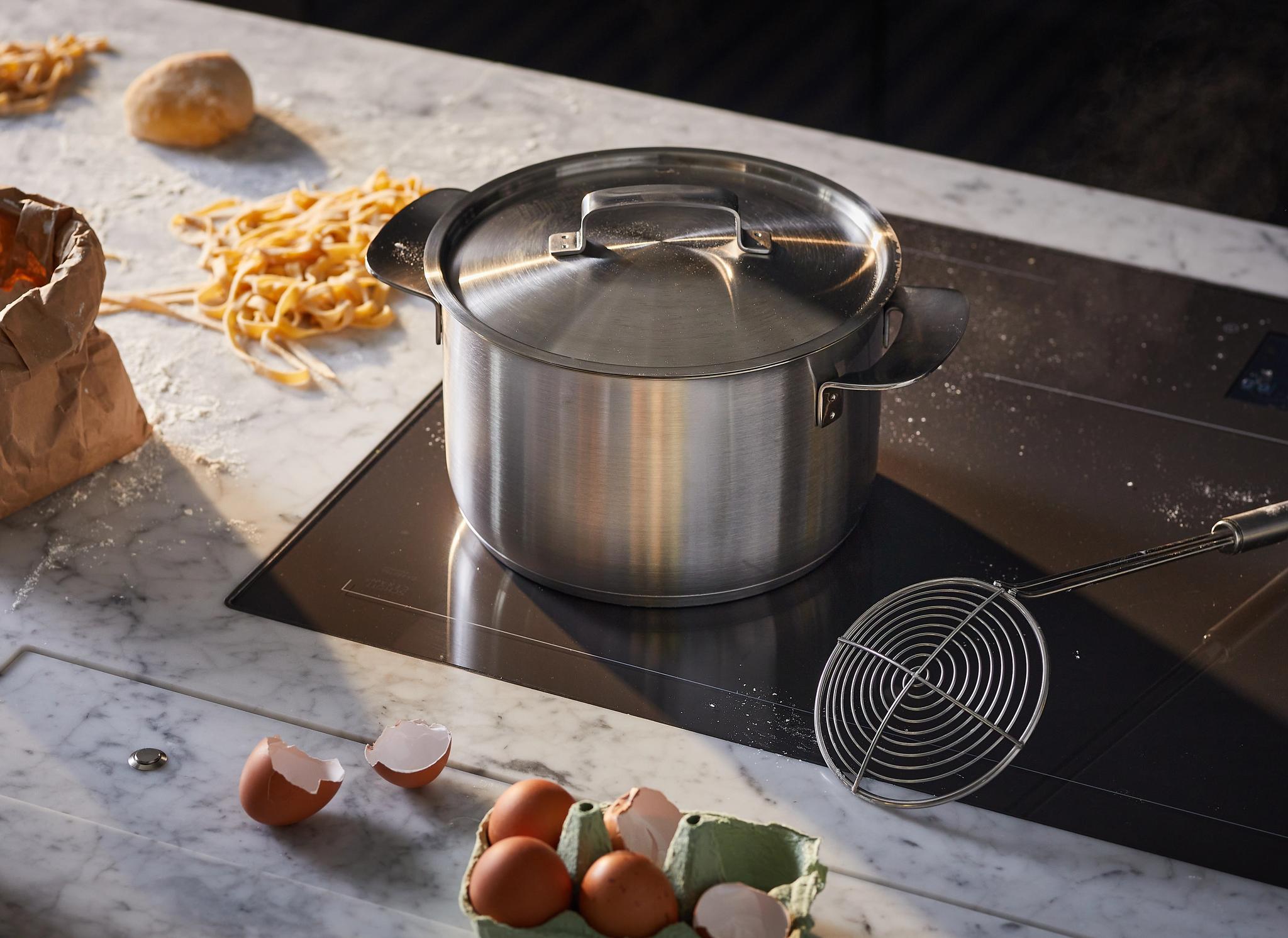 Kotitekoisen pastan ainekset hajallaan marmorisella keittiötasolla, kattila induktioliesitasolla.