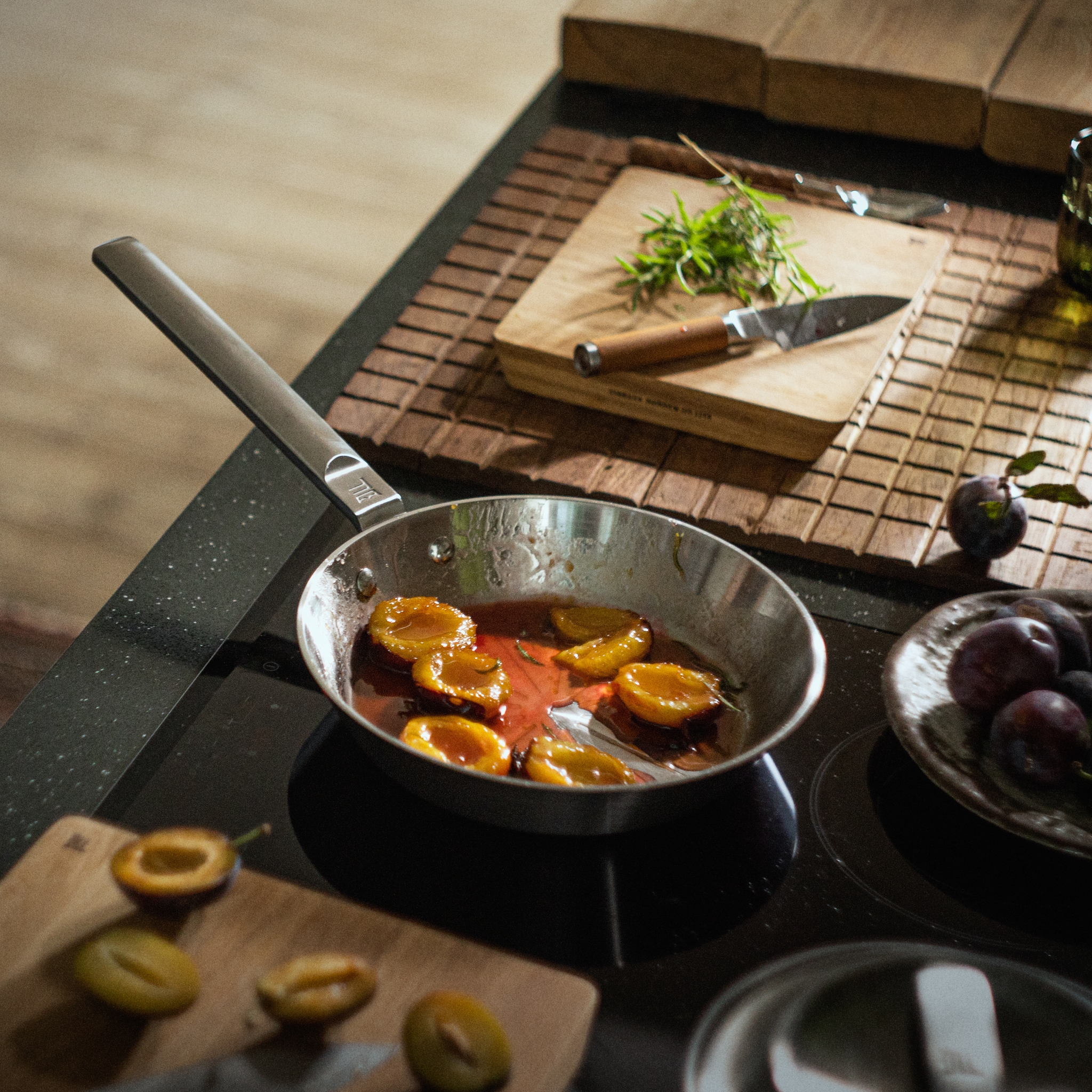 Luumunpuolikkaat karamellisoituvat pannulla liedellä tuoreiden luumujen ja rosmariinin ympäröimänä.