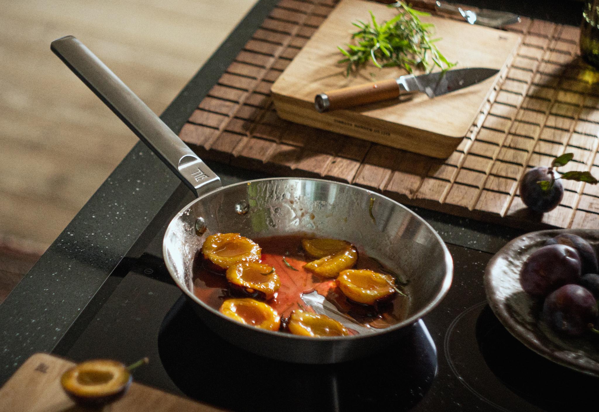 Luumunpuolikkaat karamellisoituvat pannulla liedellä tuoreiden luumujen ja rosmariinin ympäröimänä.