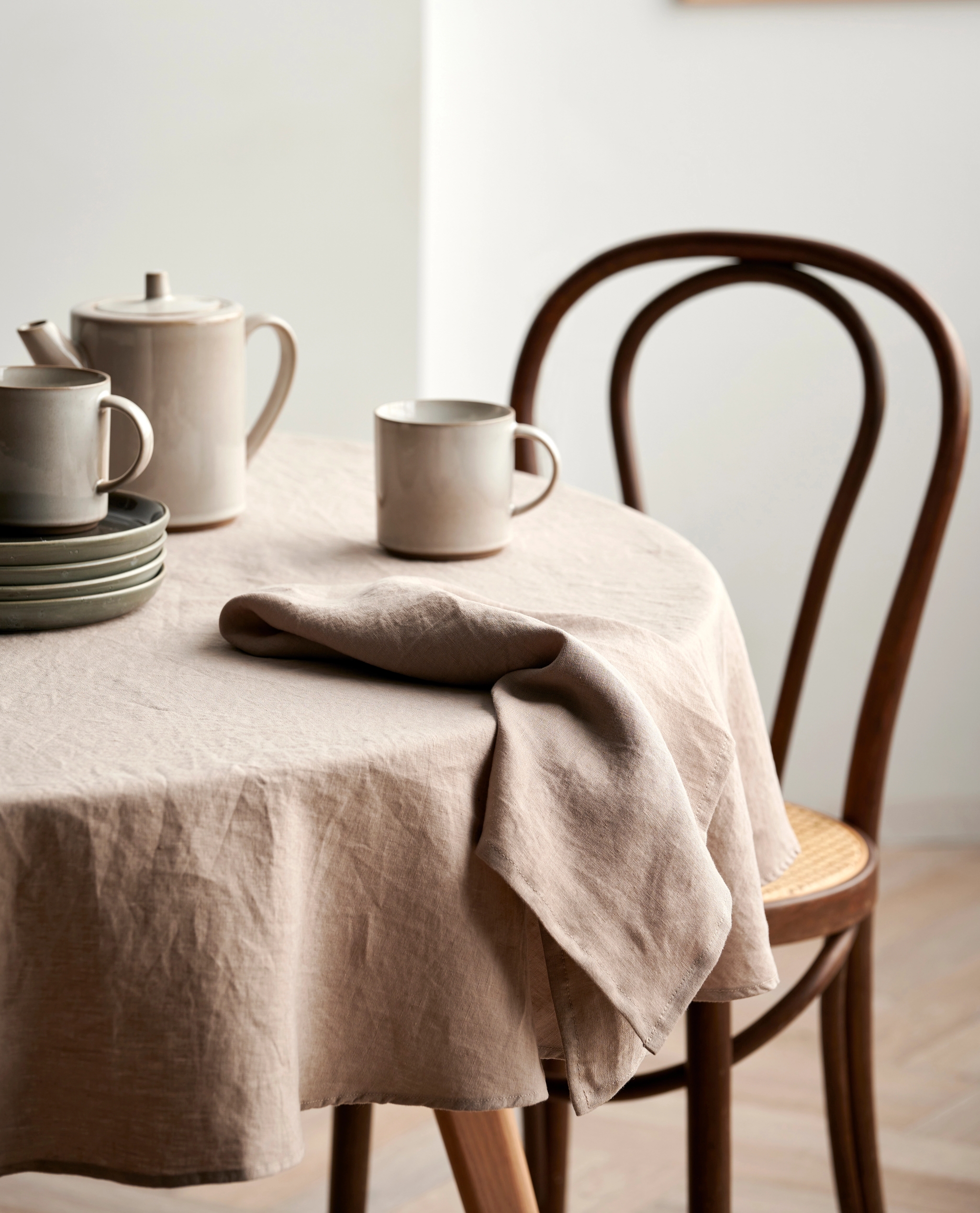 Ett runt bord täckt med en beige linneduk, dukat med stengodsservis för te och mörka tallrikar, bredvid en trästol