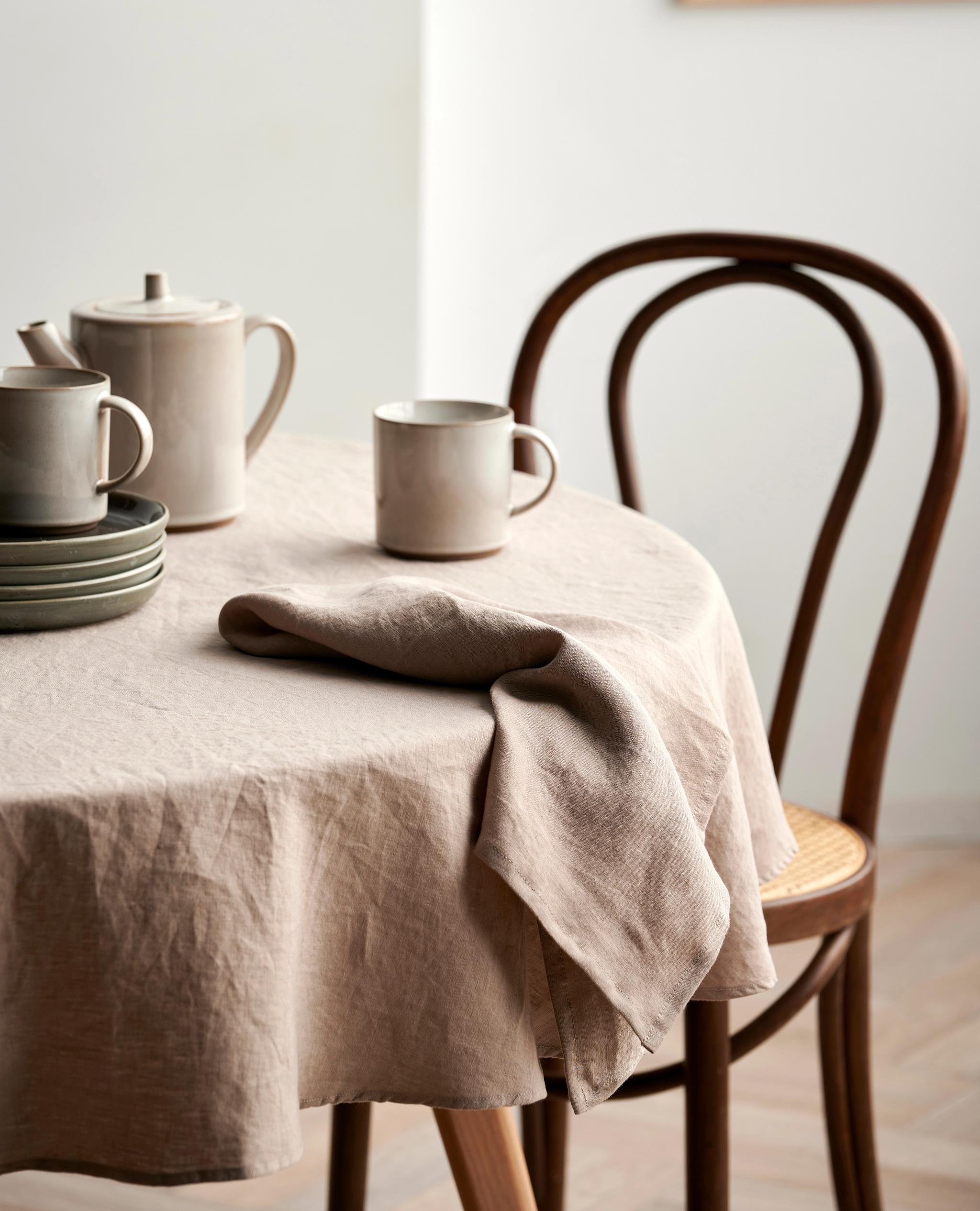 Ett runt bord täckt med en beige linneduk, dukat med stengodsservis för te och mörka tallrikar, bredvid en trästol