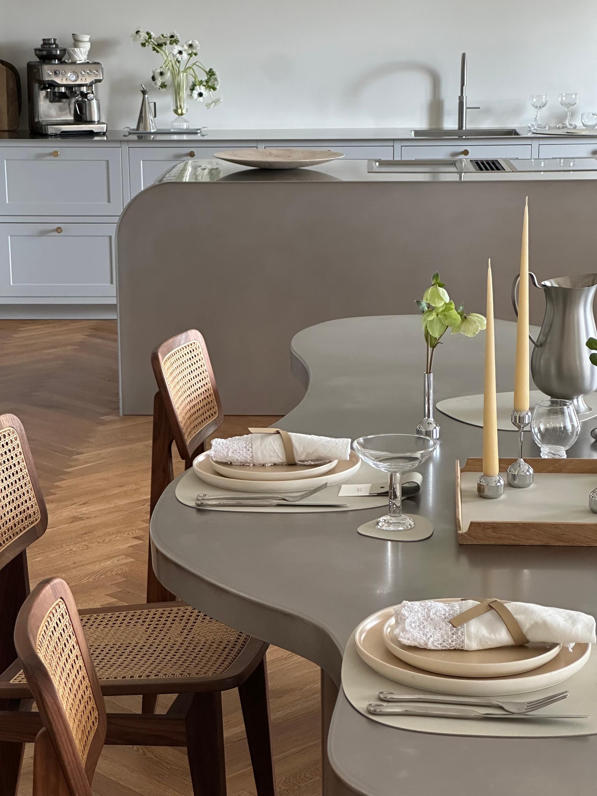 Matplats med organiskt formad bordsskiva, ljusa rottingstolar och stilren dukning i beige toner, med eleganta glas, linne-servetter och smala ljus i ett modernt kök med grå köksö i bakgrunden.