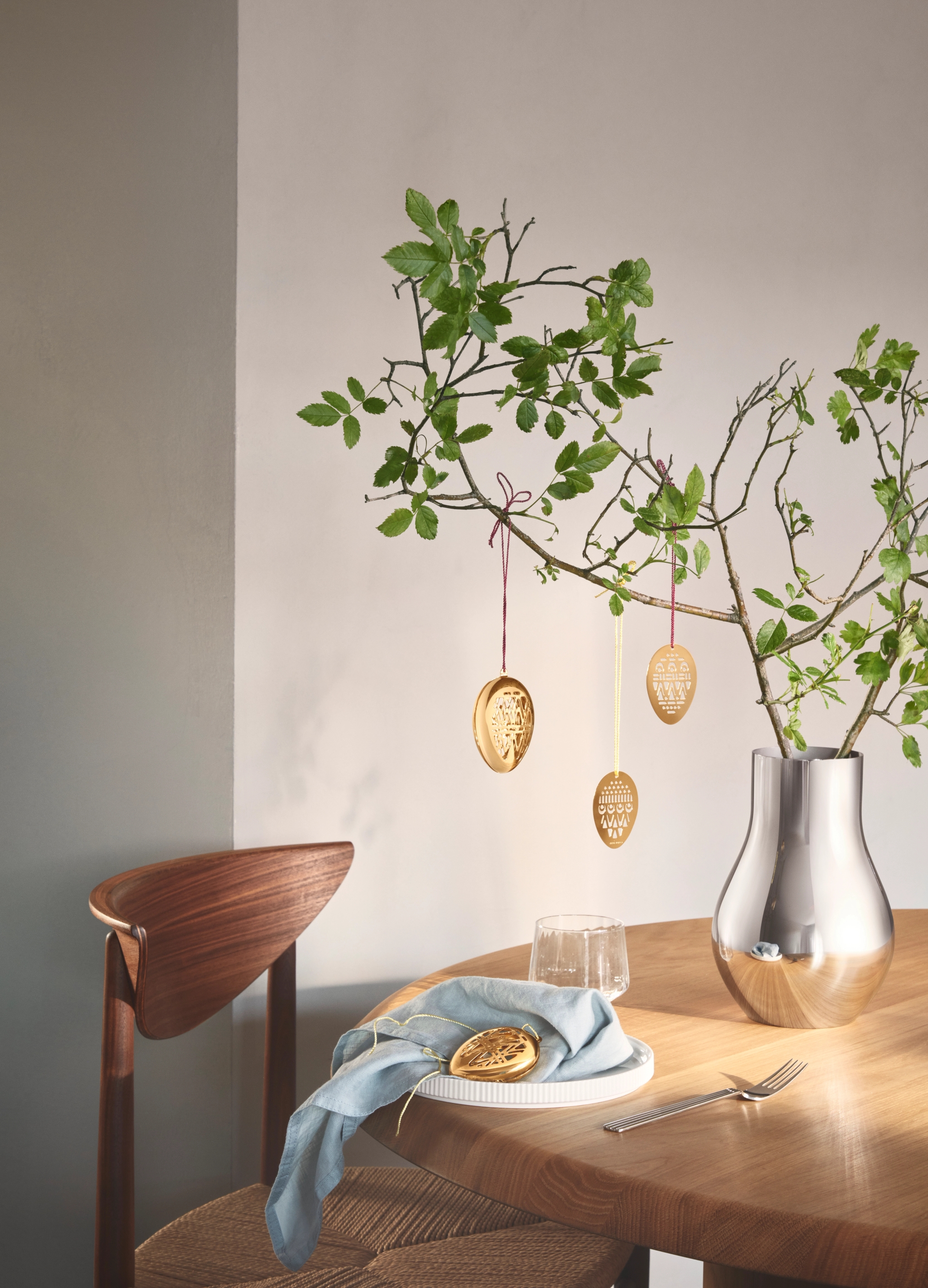 Påskdukning med gyllene äggdekorationer hängande från gröna grenar i en silvervas och ett dekorativt ägg på ett fat.