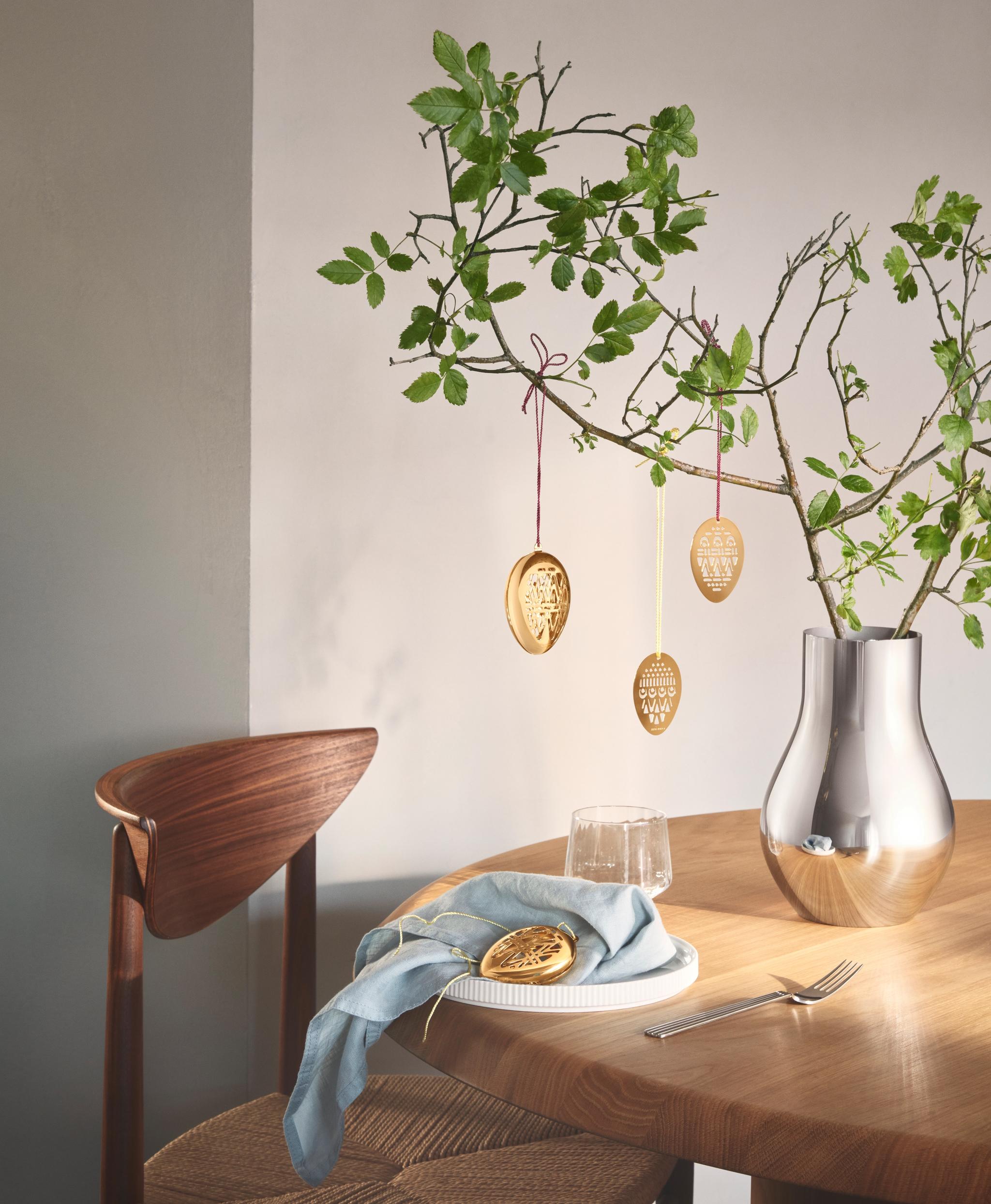 Påskdukning med gyllene äggdekorationer hängande från gröna grenar i en silvervas och ett dekorativt ägg på ett fat.