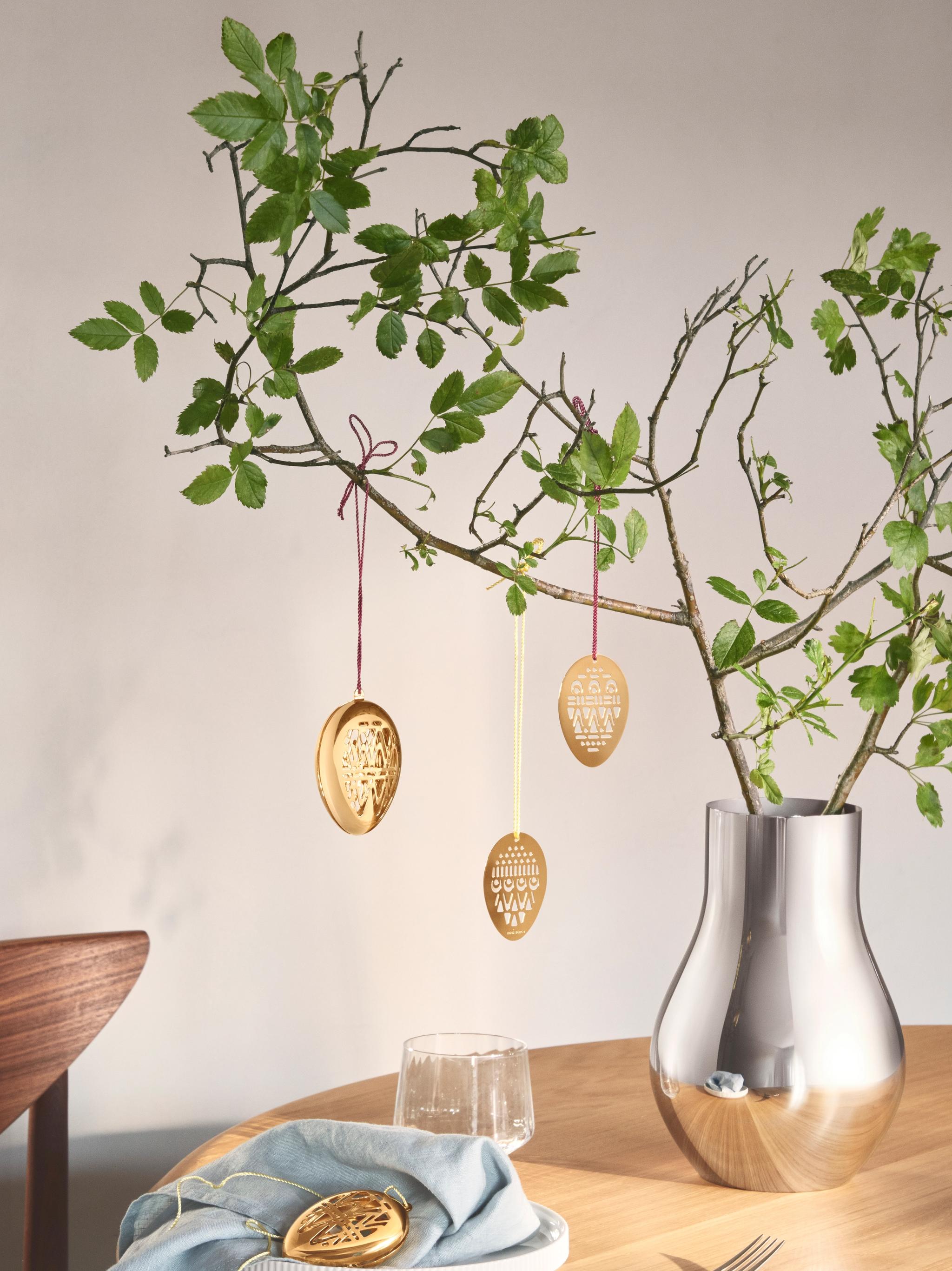 En sølvvase holder løvrike grener pyntet med gullfargede påskeeggpynt på et trebord.