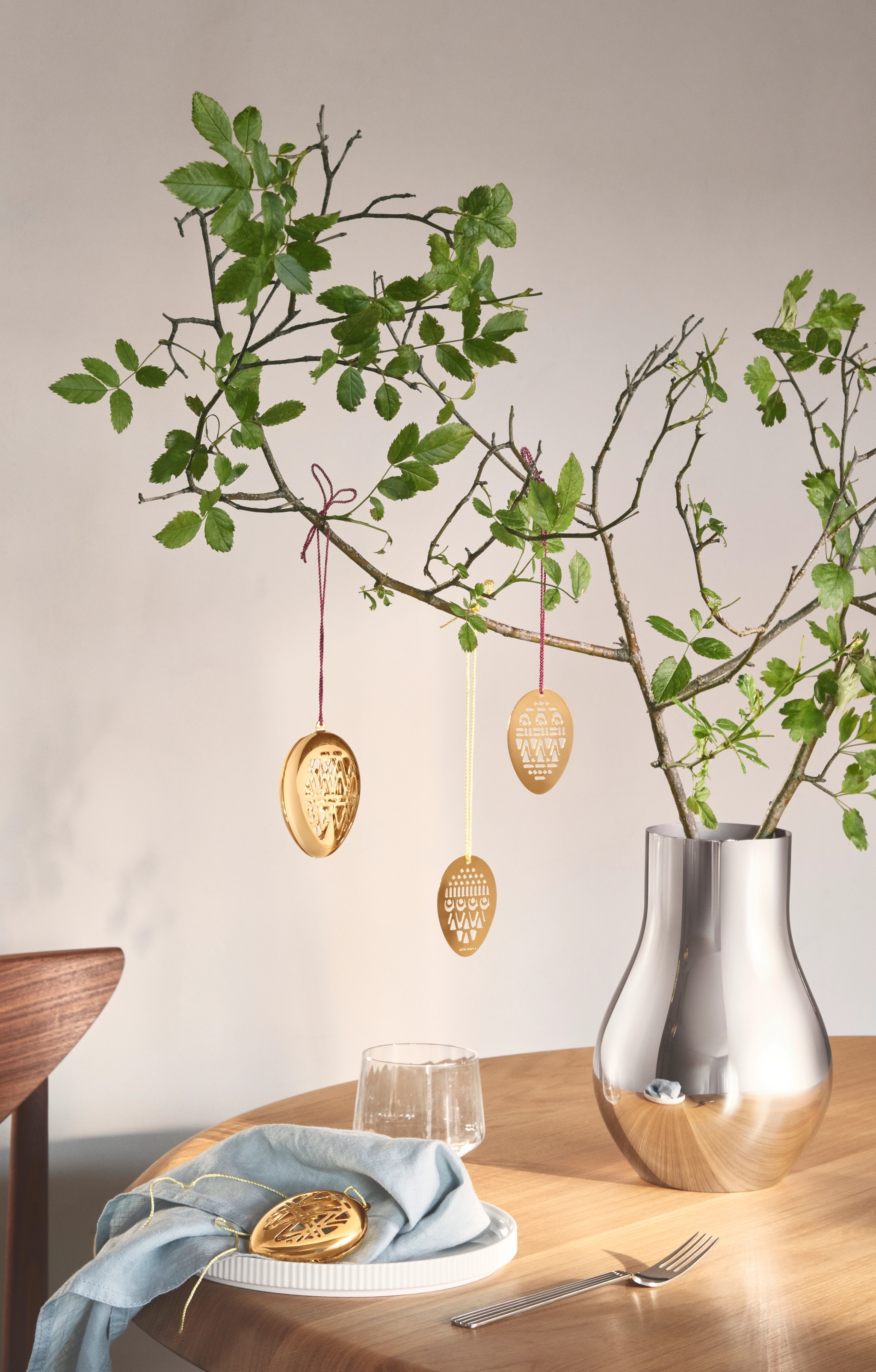 En sølvvase holder løvrike grener pyntet med gullfargede påskeeggpynt på et trebord.