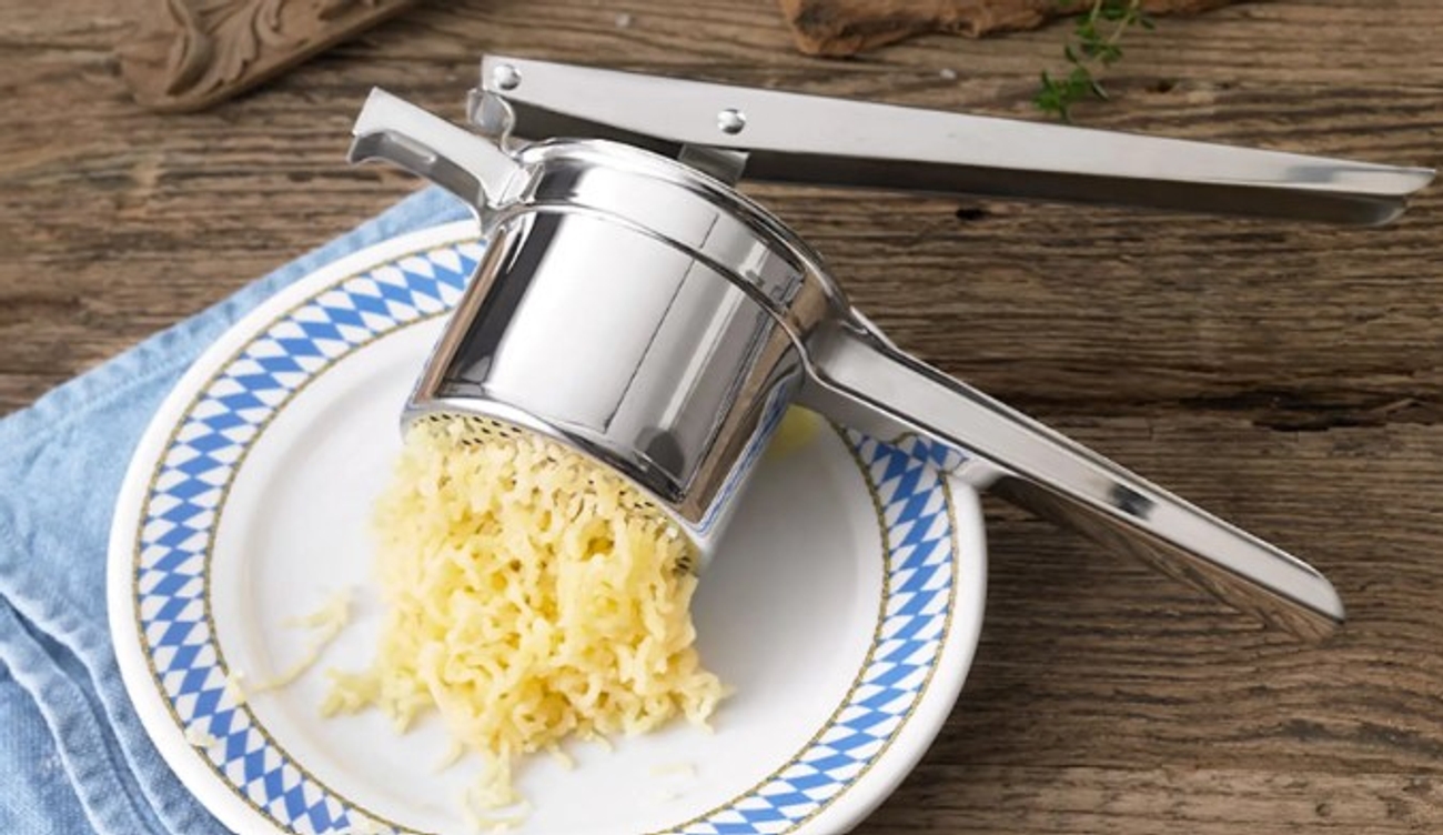 Potatispressar i silver som pressar potatis på vit tallrik med blått mönster runt kanten 