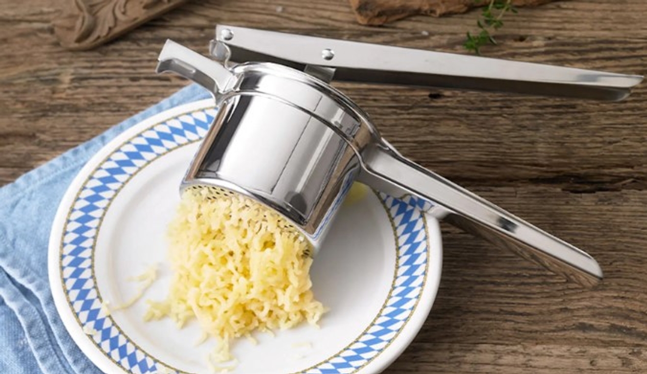 Potatispressar i silver som pressar potatis på vit tallrik med blått mönster runt kanten 
