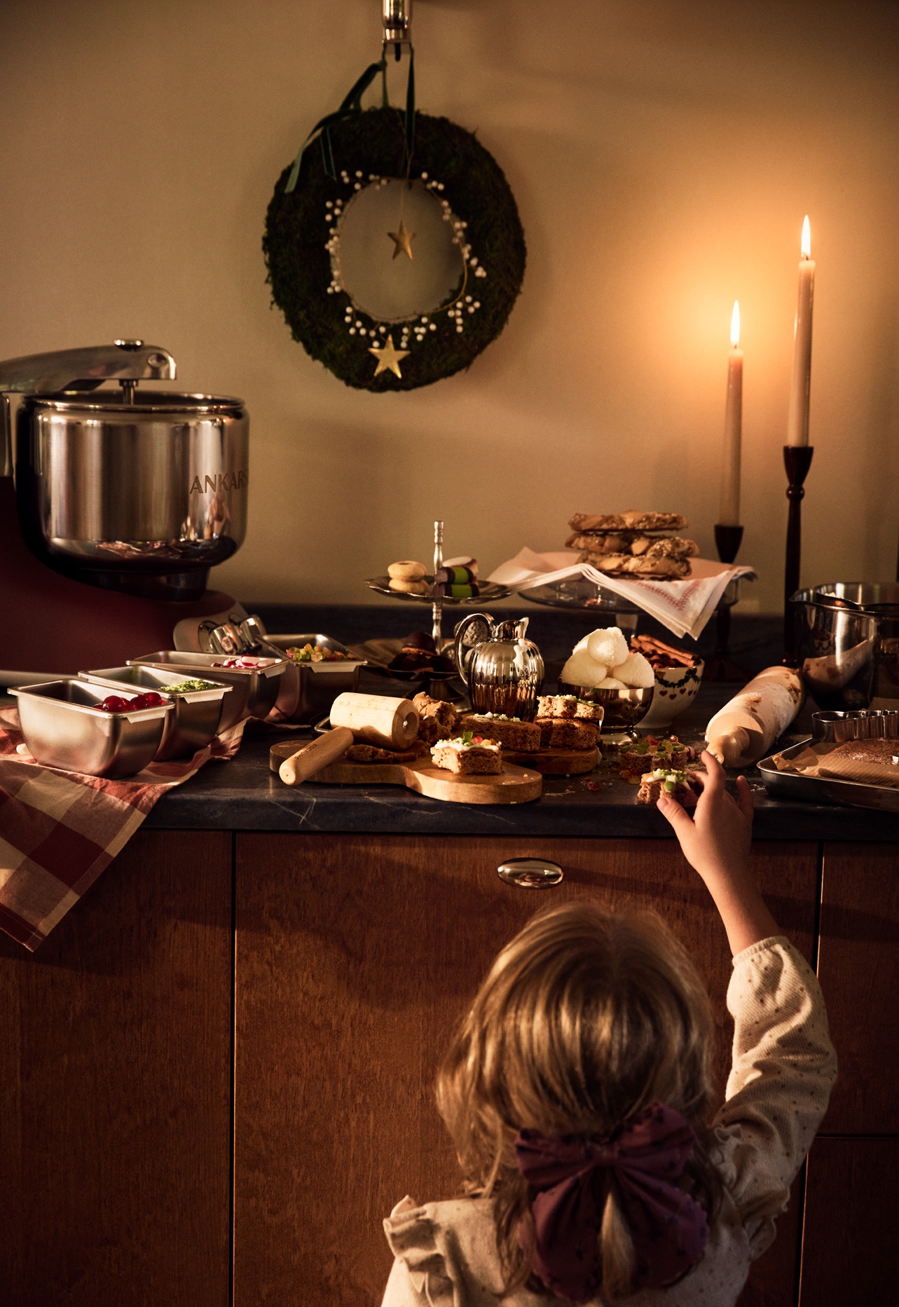 Ett barn sträcker sig efter julgodis på ett stämningsfullt köksbord med bakning, tända ljus och juldekorationer.