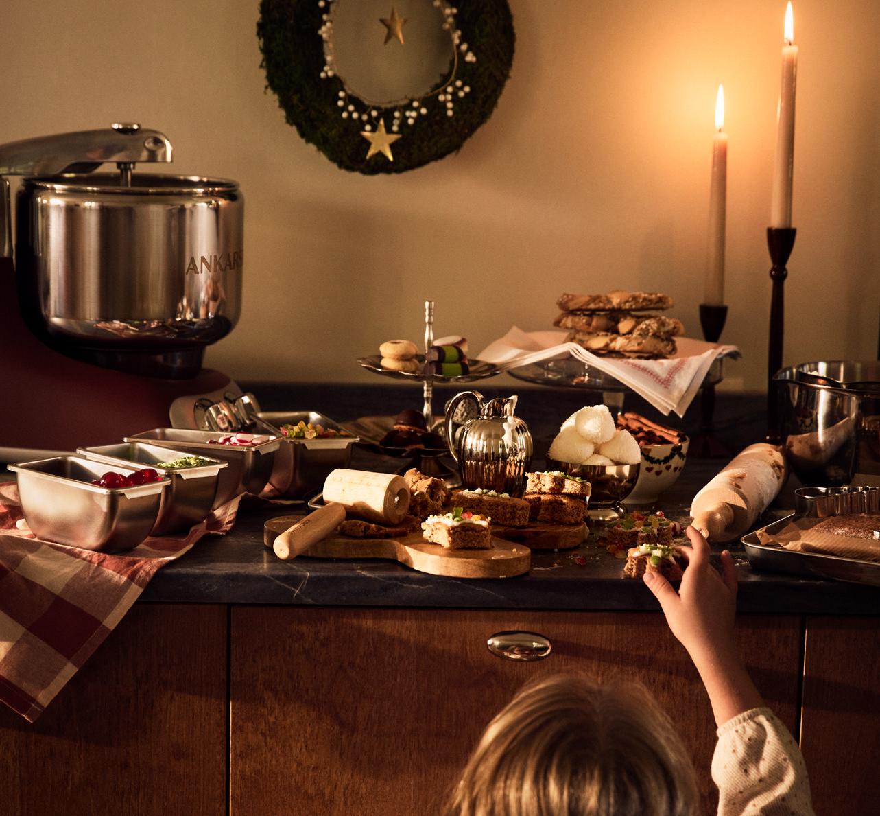 Ett barn sträcker sig efter julgodis på ett stämningsfullt köksbord med bakning, tända ljus och juldekorationer.