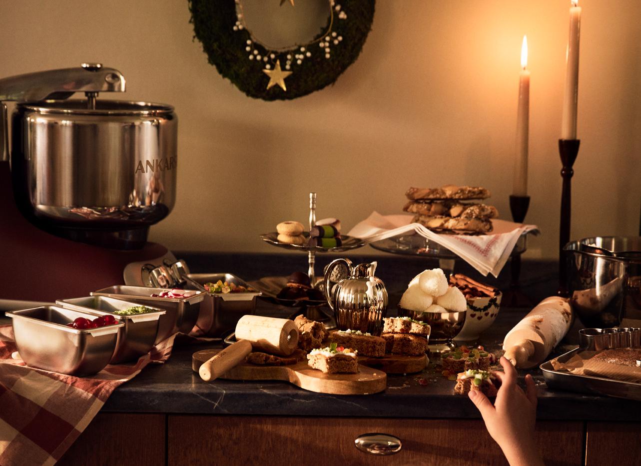 Ett barn sträcker sig efter julgodis på ett stämningsfullt köksbord med bakning, tända ljus och juldekorationer.