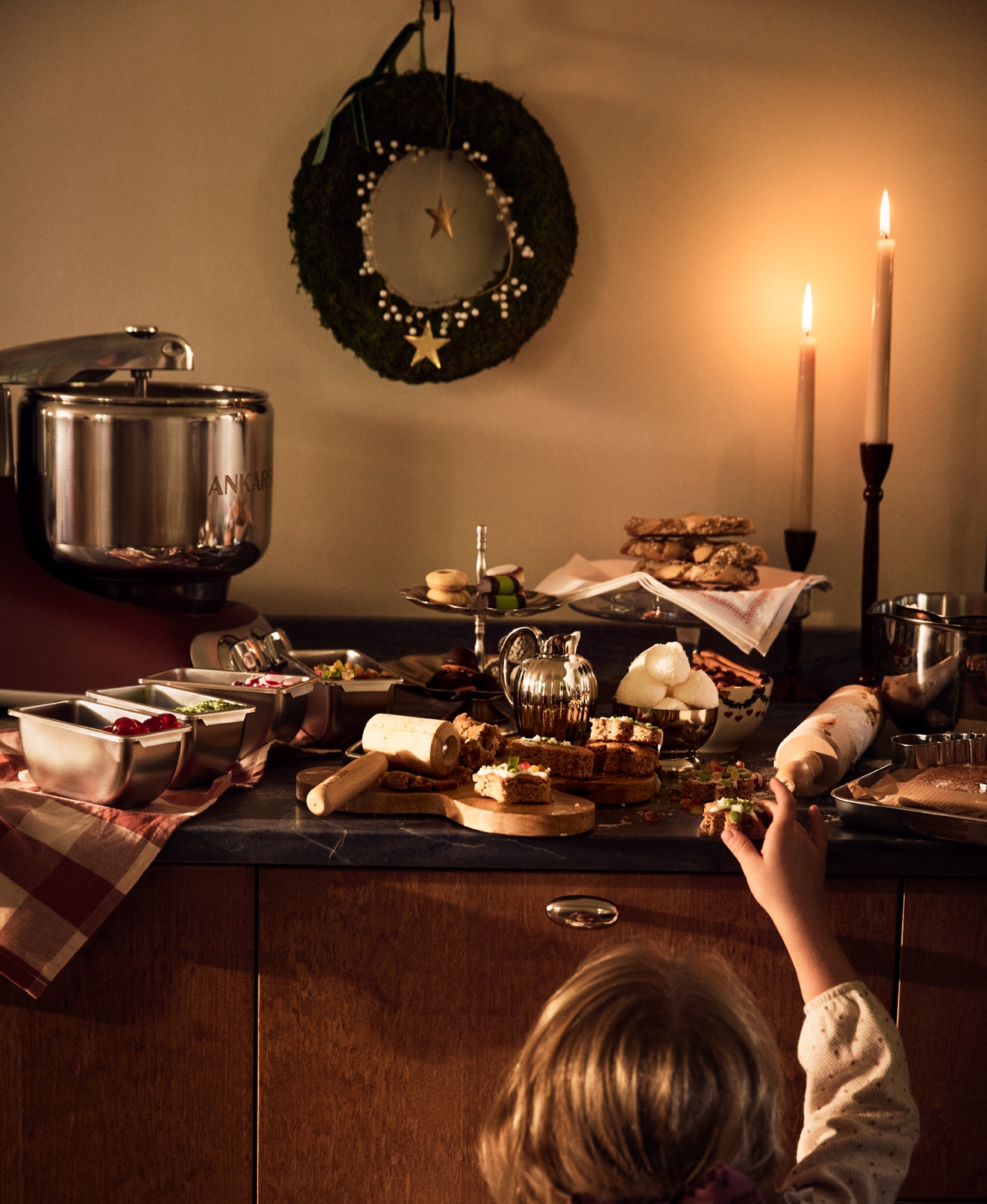 Ett barn sträcker sig efter julgodis på ett stämningsfullt köksbord med bakning, tända ljus och juldekorationer.