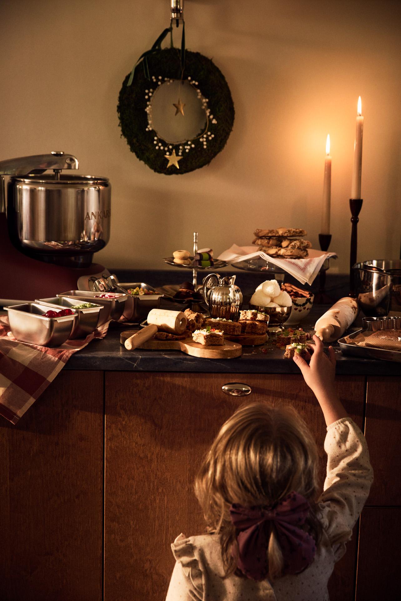 Ett barn sträcker sig efter julgodis på ett stämningsfullt köksbord med bakning, tända ljus och juldekorationer.