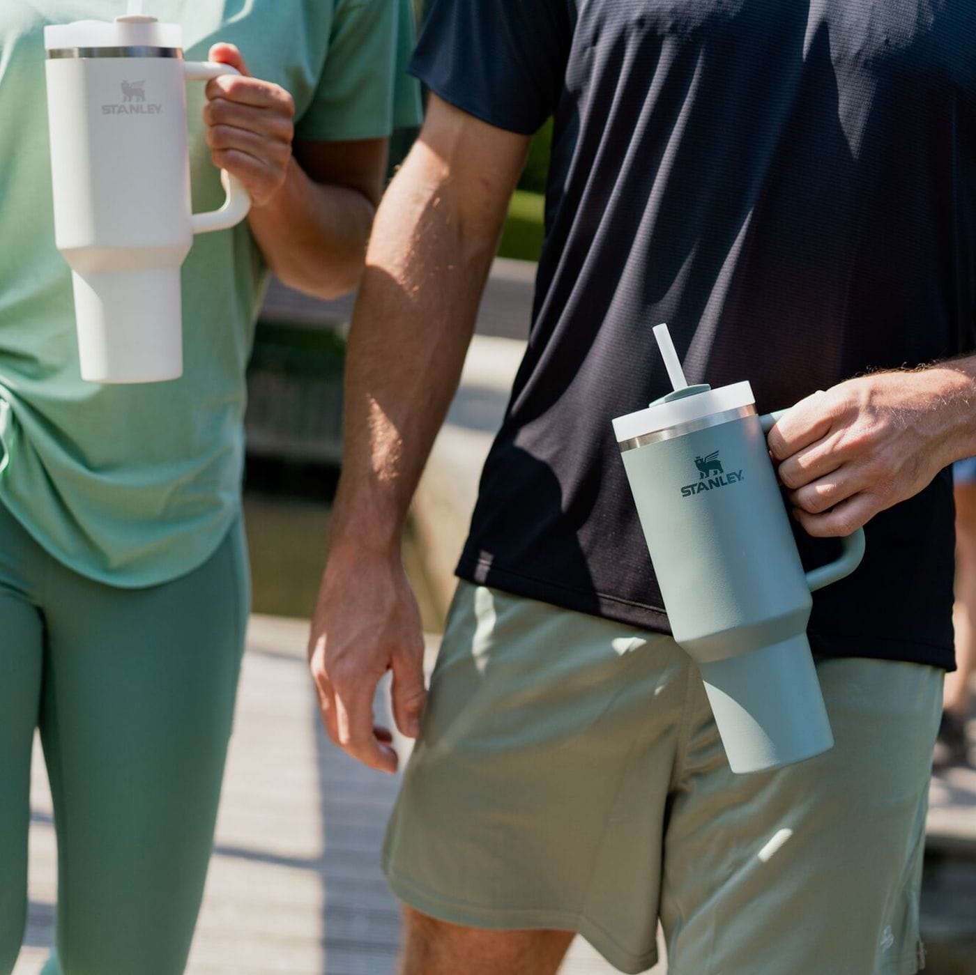 Två personer i träningskläder håller varsin Stanley-tumbler, en vit och en grön.