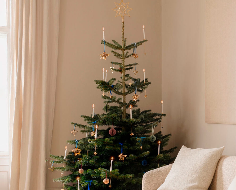 A decorated Christmas tree with a star topper, lit candles, and wrapped gifts at its base, next to a cream sofa.