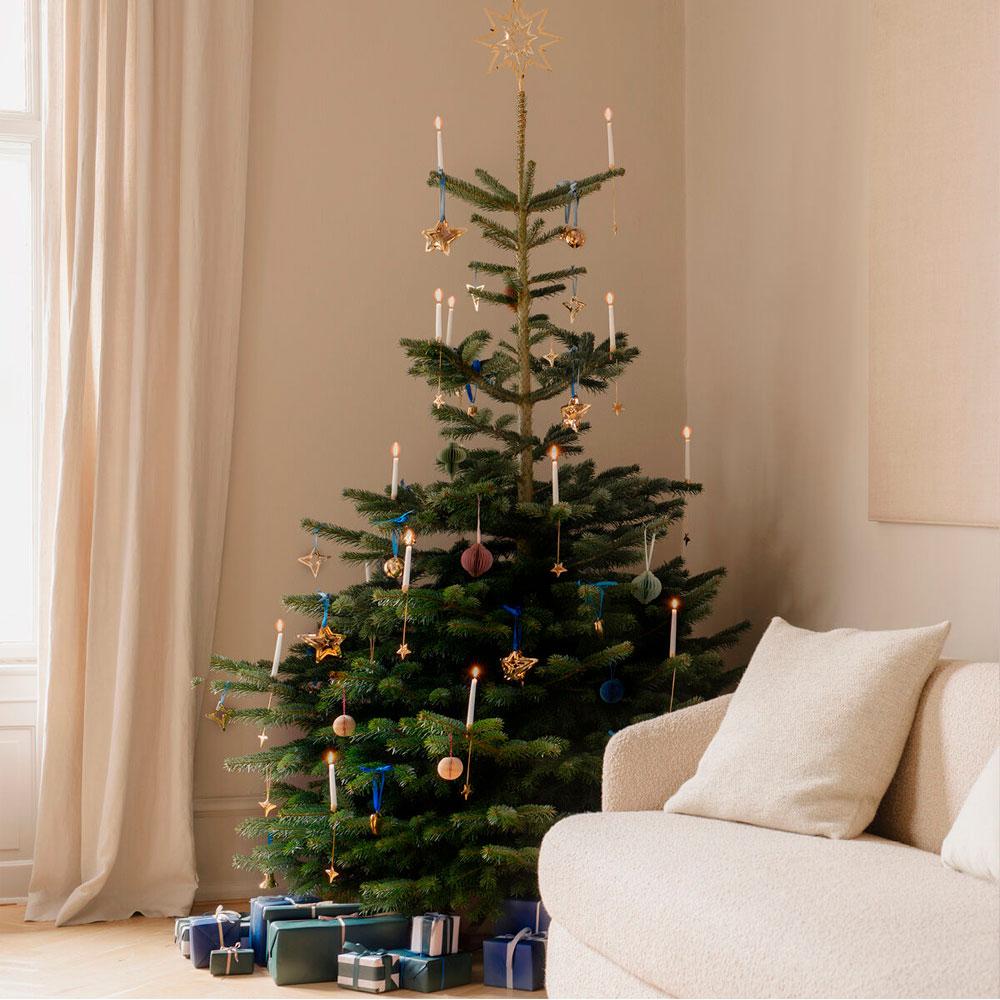 A Christmas tree with lit candles, a gold star topper, and ornaments, with gifts at its base, next to a light sofa.