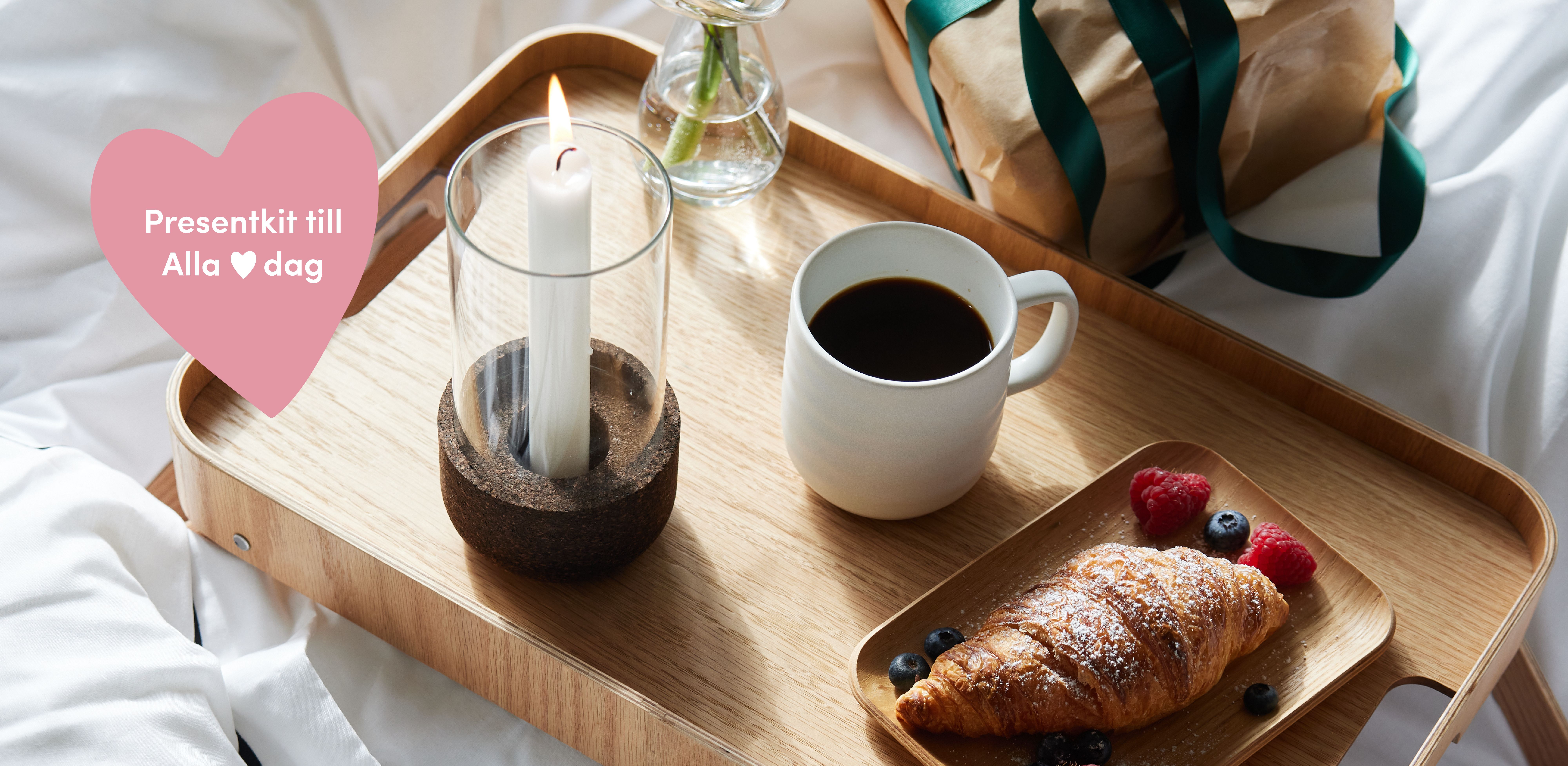 Breakfast in bed on a wooden tray with a croissant, coffee, and lit candle, next to a wrapped gift. A pink heart overlay reads 'Presentkit till Alla ❤️ dag'.