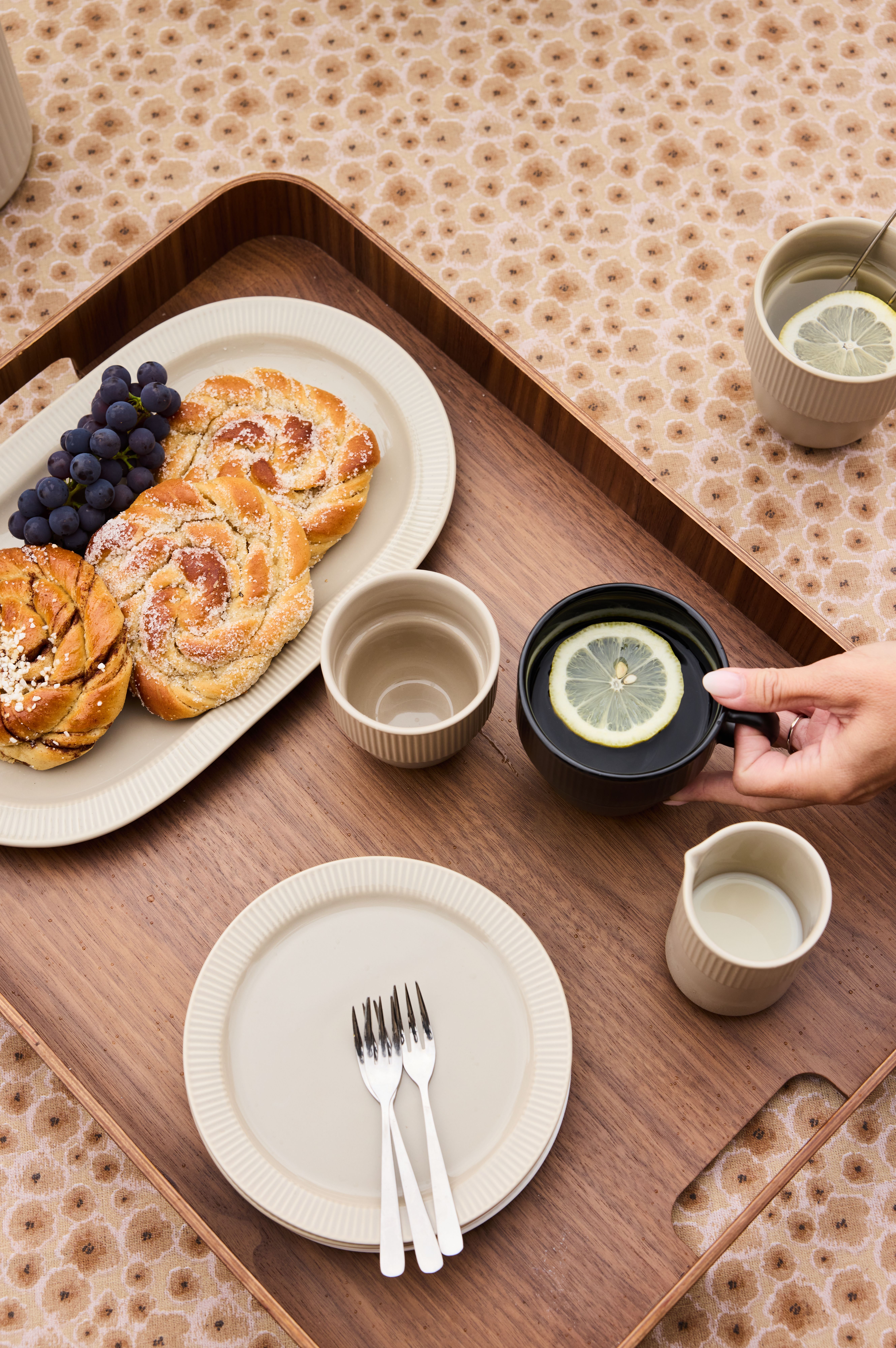 En träbricka med bakverk, vindruvor, te med citron och grädde på en blommig duk, med en hand som håller i en mörk mugg