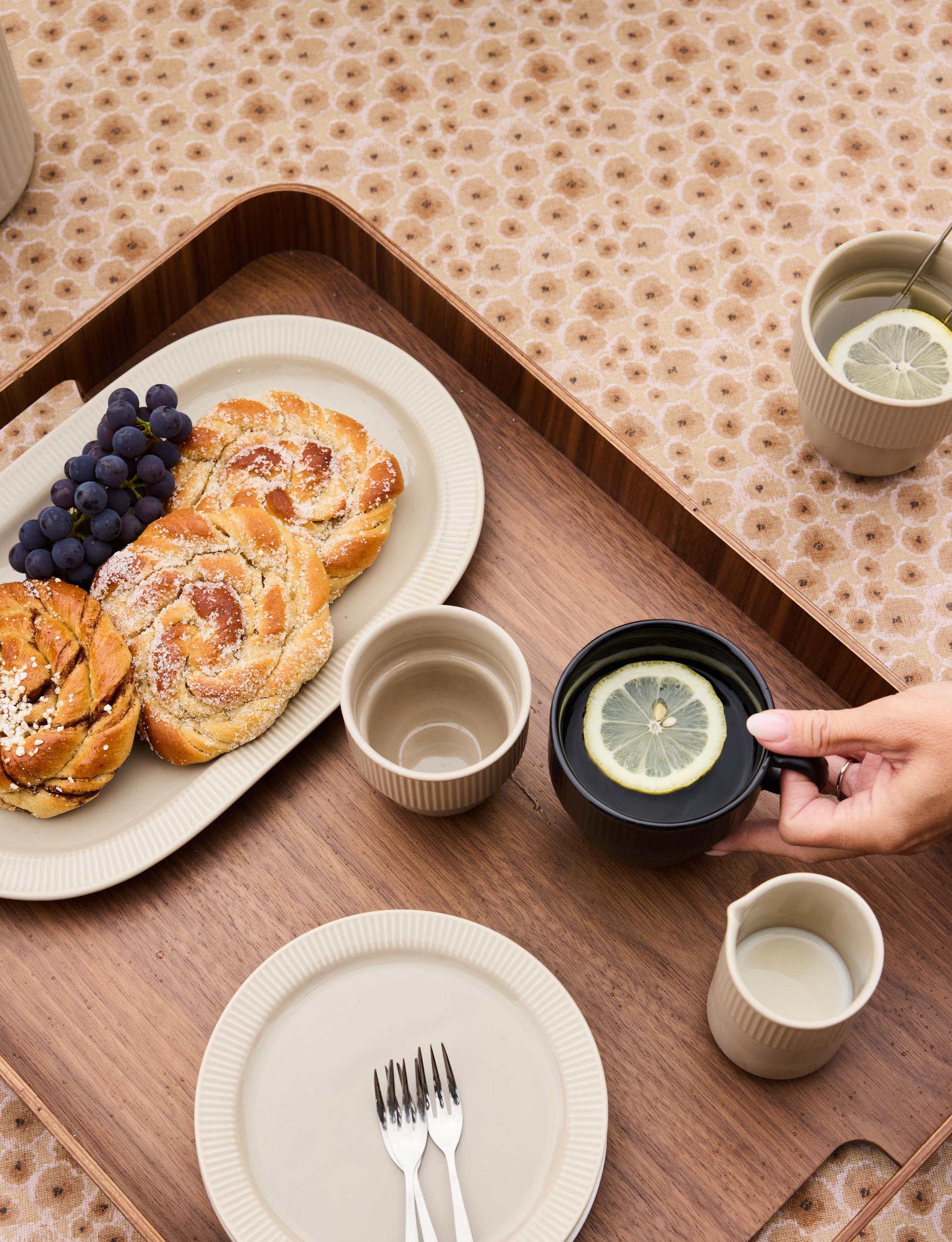 En träbricka med bakverk, vindruvor, te med citron och grädde på en blommig duk, med en hand som håller i en mörk mugg
