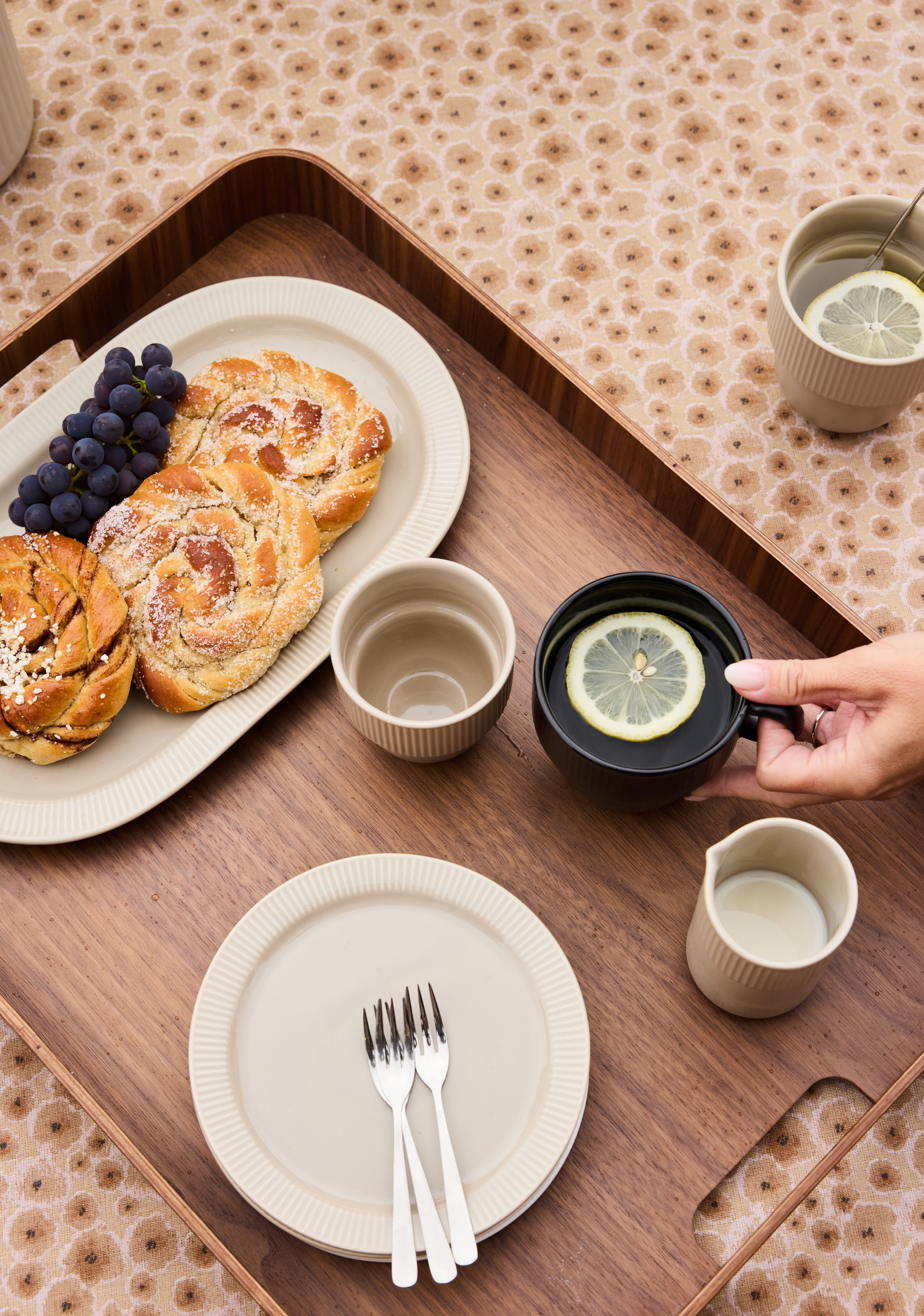 En träbricka med bakverk, vindruvor, te med citron och grädde på en blommig duk, med en hand som håller i en mörk mugg