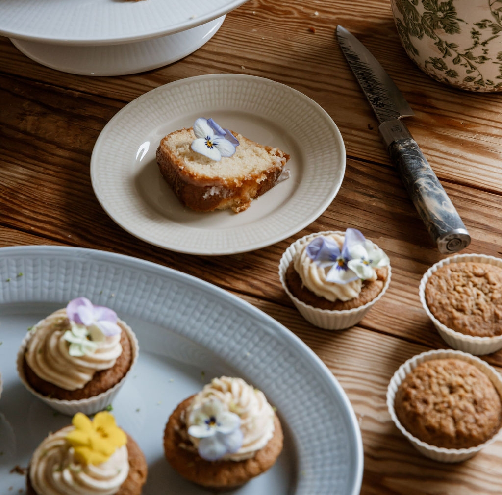 Saftig formkake og cupcakes pyntet med spiselige blomster står på et rustikt trebord ved siden av en blomsterpotte.